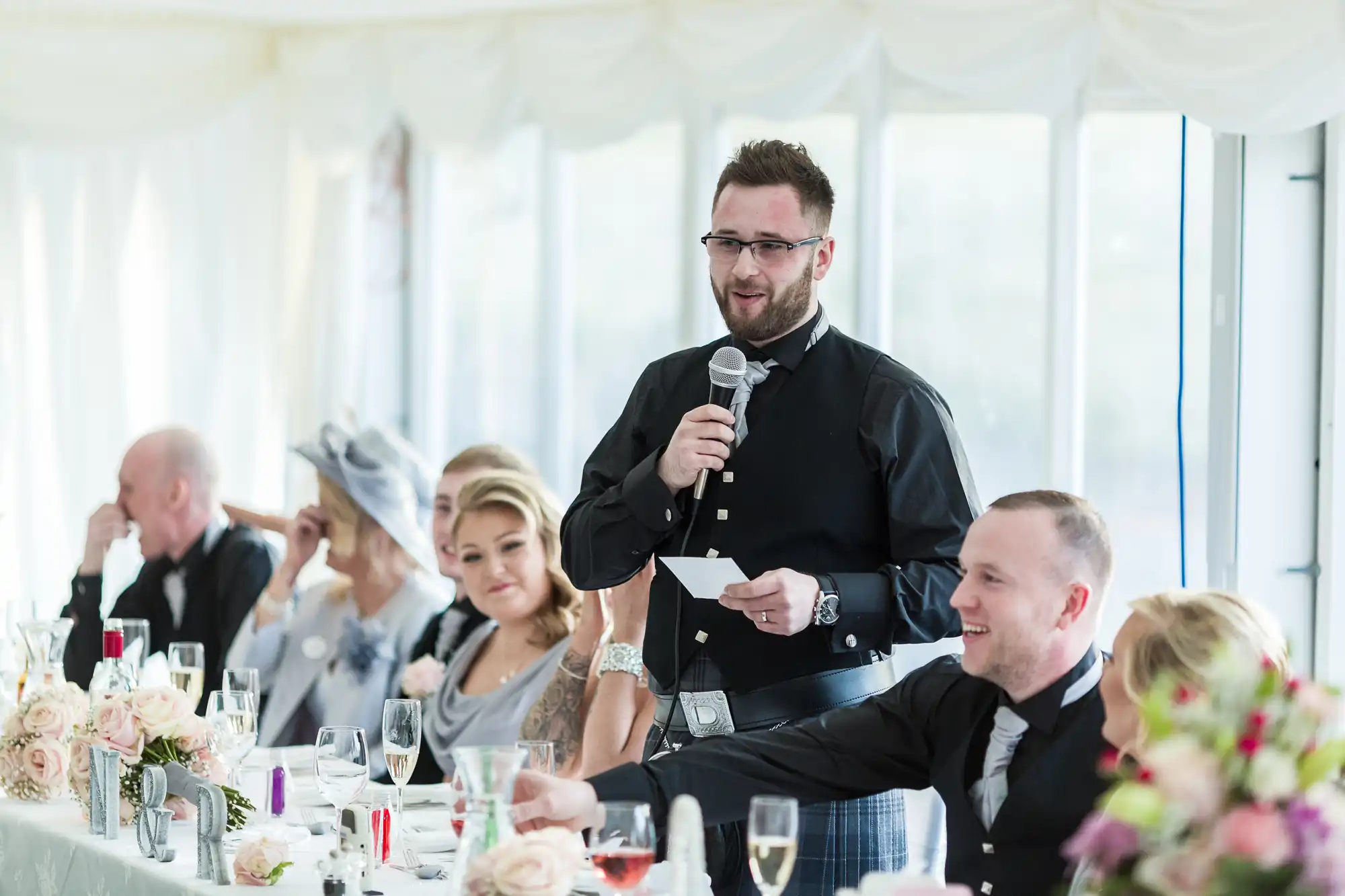 A man in a black shirt and kilt stands holding a microphone and a note, speaking at a long, elegantly decorated table. Seated guests, some dressed formally, listen attentively. The room is bright with large windows and floral centrepieces on the table.