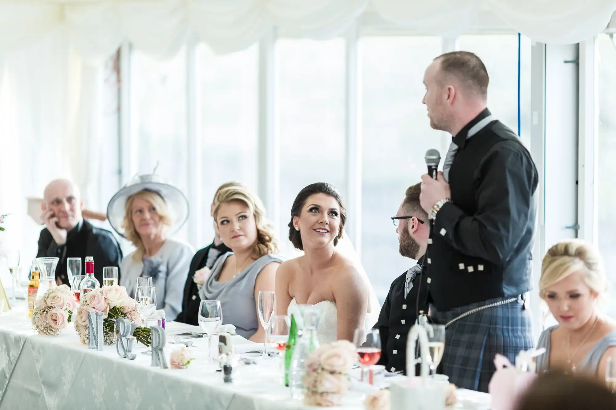 A bride in a strapless dress smiles at a man in a kilt giving a speech with a microphone at a decorated wedding reception table. Guests in formal attire sit beside them, surrounded by flowers, glassware, and white table linen in a bright room.