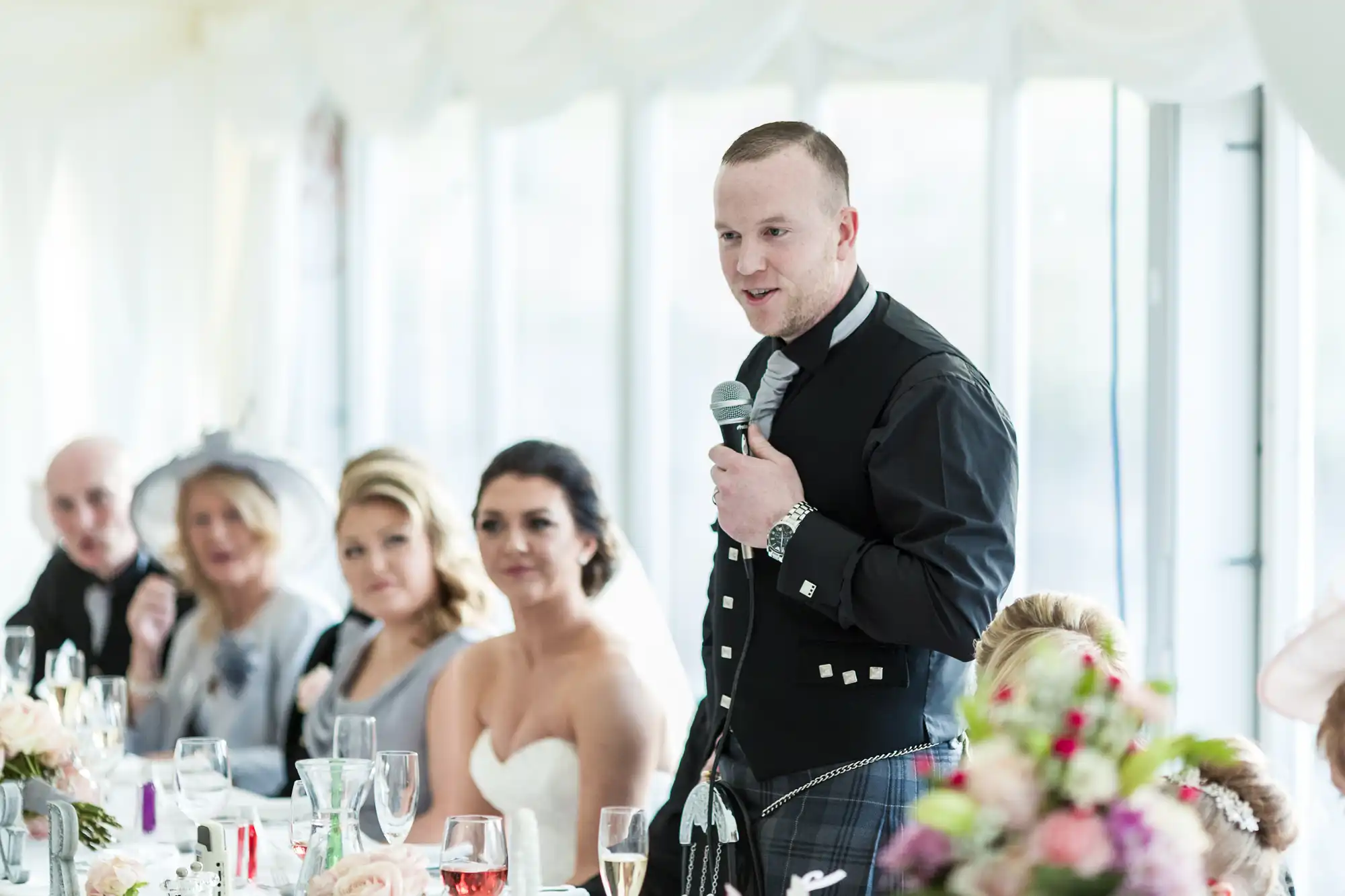 A man in a black kilt outfit stands holding a microphone, giving a speech at a wedding reception. Guests, including a bride in a strapless dress and others in formal attire, are seated at a decorated table with flowers and glasses.