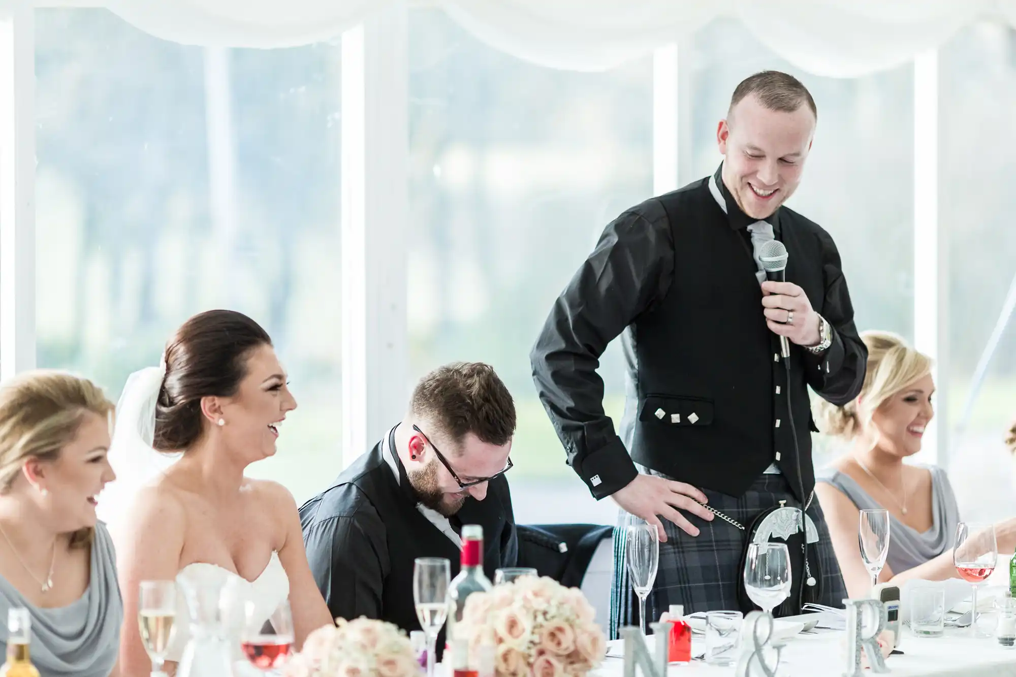 A man in a black jacket and kilt stands smiling with a microphone, giving a speech at a wedding reception. Four guests in formal attire, including a bride in white, sit at a table decorated with flowers and wine glasses, laughing and smiling.