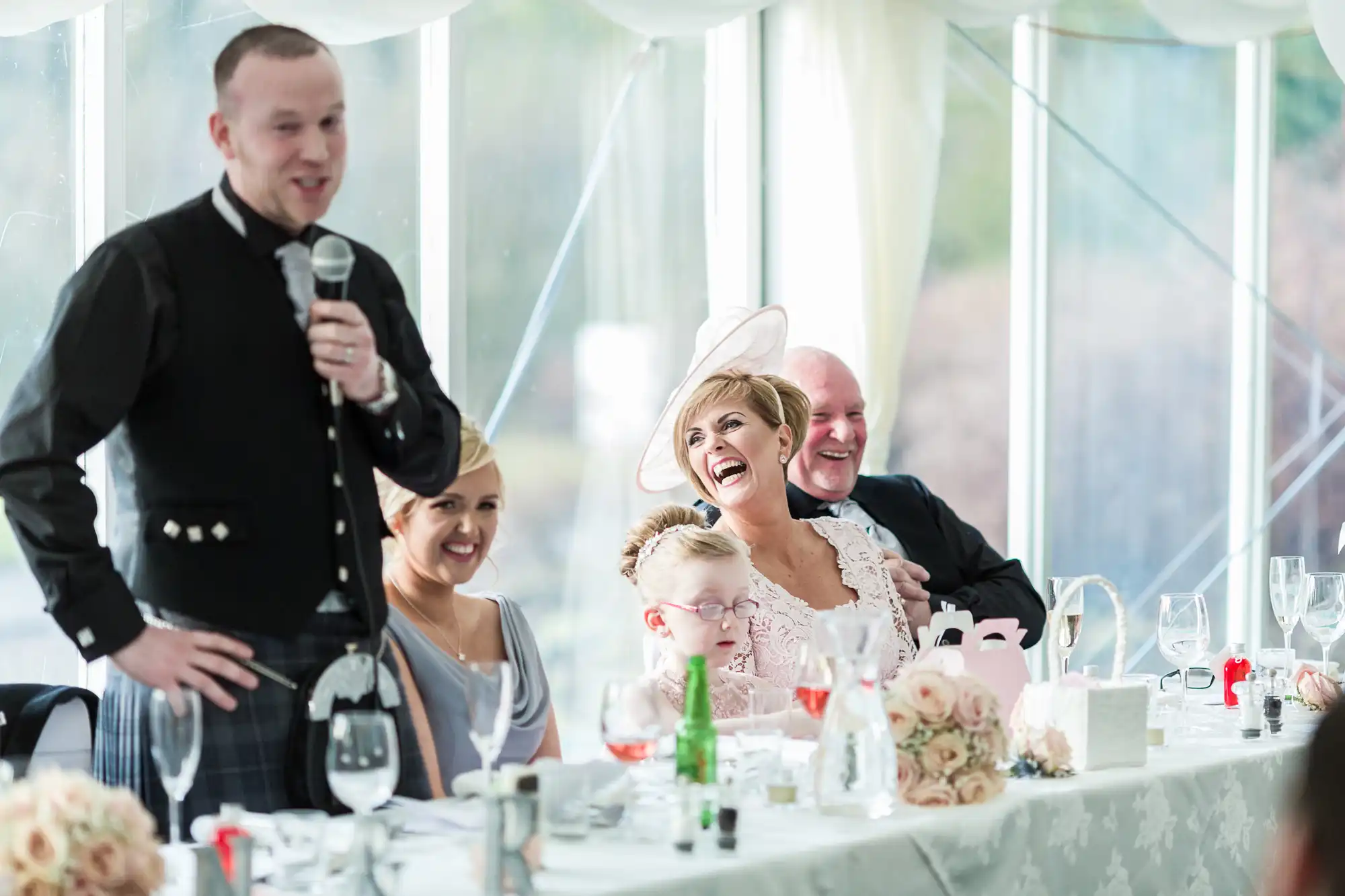 A man in traditional Scottish attire speaks into a microphone at a wedding reception. Seated guests, including a smiling bride in a hat, a child with glasses, and other adults, listen and laugh at a table decorated with flowers and wine glasses.