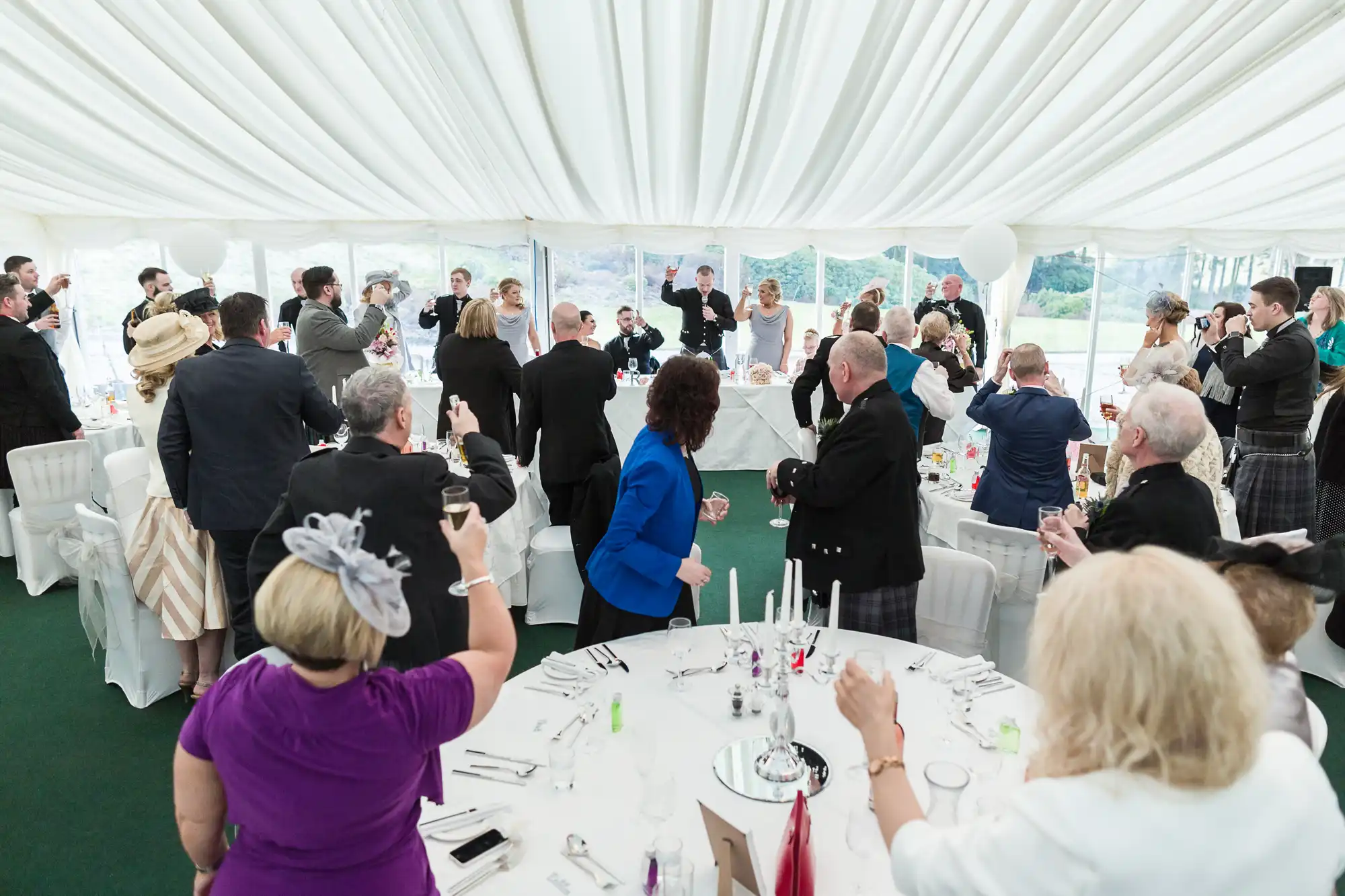Wedding guests in formal attire stand around decorated tables inside a white marquee, raising glasses in a toast towards a top table where the bridal couple are seated. The atmosphere is festive, with white drapery and natural light filling the marquee.