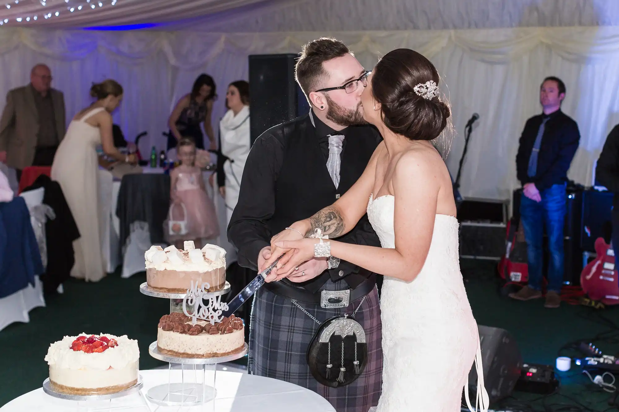 A bride and groom kiss while cutting a tiered wedding cake together at a reception. The bride wears a white strapless gown and the groom wears a black jacket and tartan kilt. Guests and musicians are visible in the background under a white marquee with fairy lights.