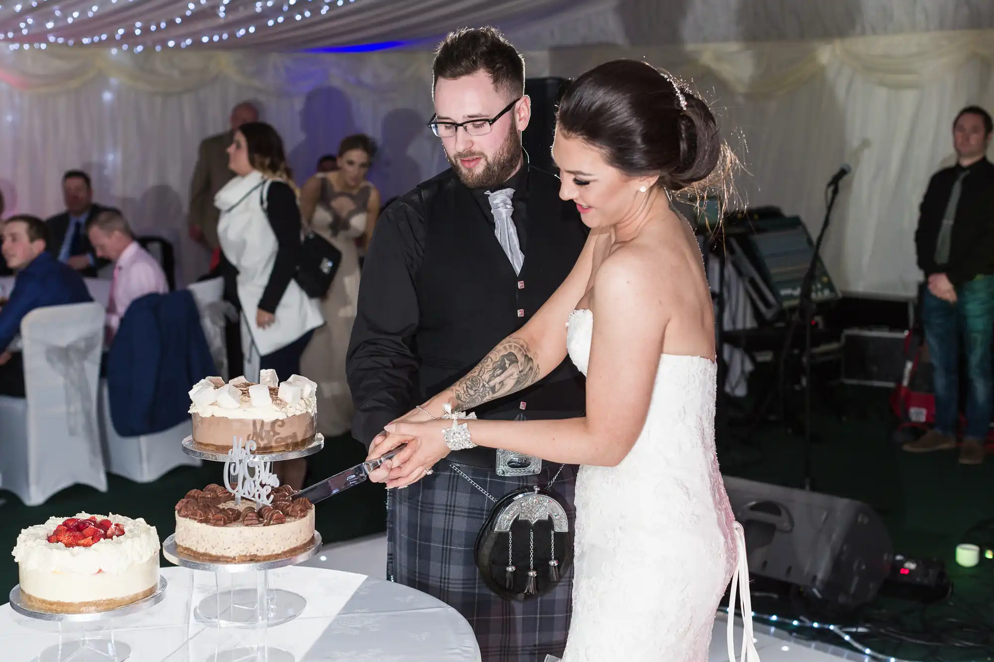 A bride in a white strapless dress and a groom in a kilt cut a two-tiered wedding cake together. They are indoors, surrounded by seated and standing guests, with white drapery and fairy lights overhead. Two more cakes are on the table.