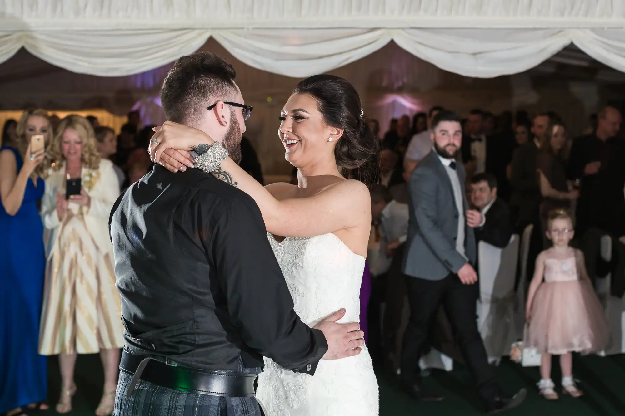 A bride in a white dress and a groom in a black shirt dance closely and smile at each other, surrounded by wedding guests. People in formal attire watch, some taking photos, while a young girl in a pink dress stands nearby. The setting is a decorated indoor venue.