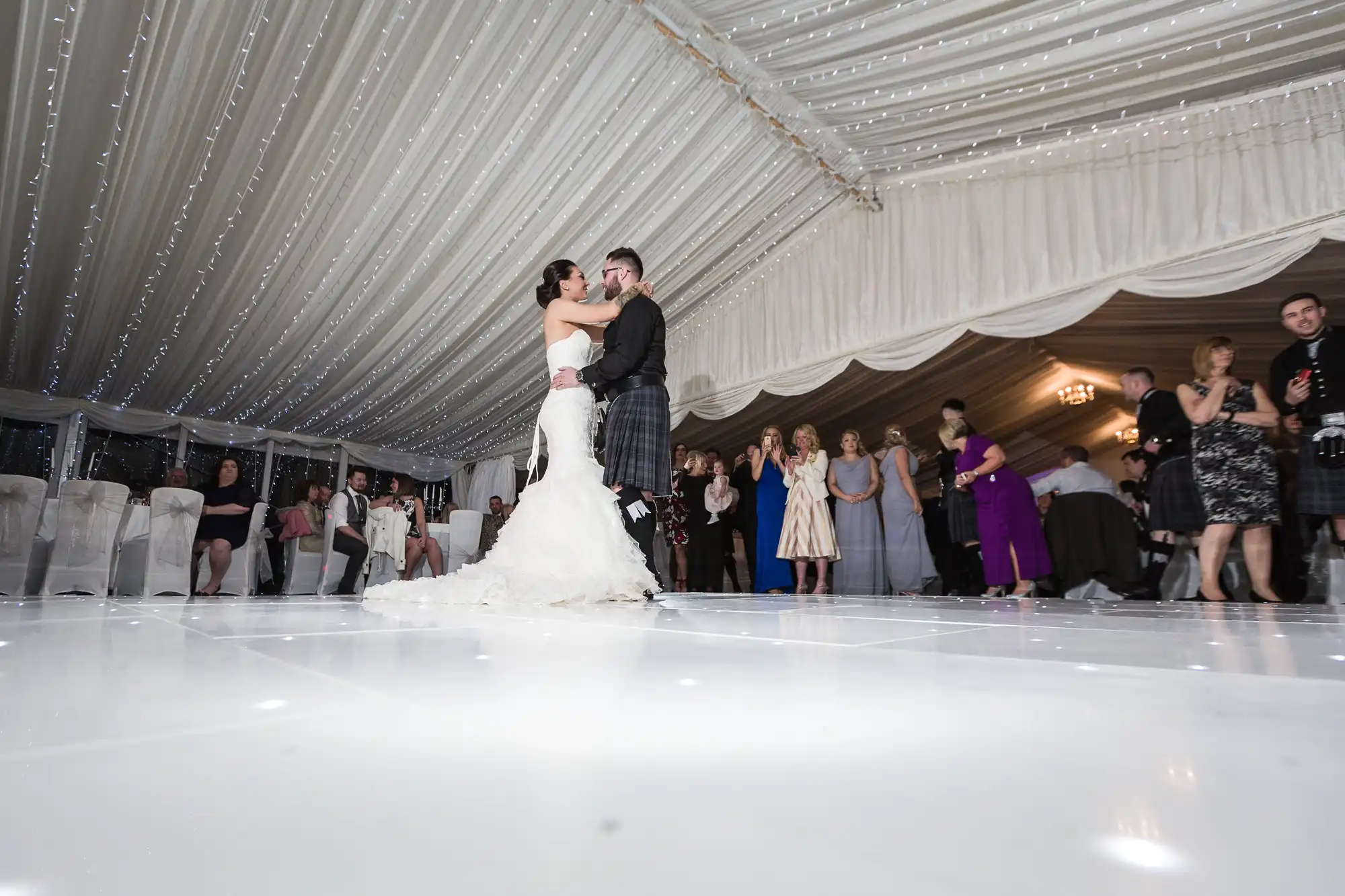 A bride in a white dress and a groom in a kilt share a dance on a glossy white floor under a draped, fairy-lit marquee, while guests seated and standing in formal attire watch and smile in the background.