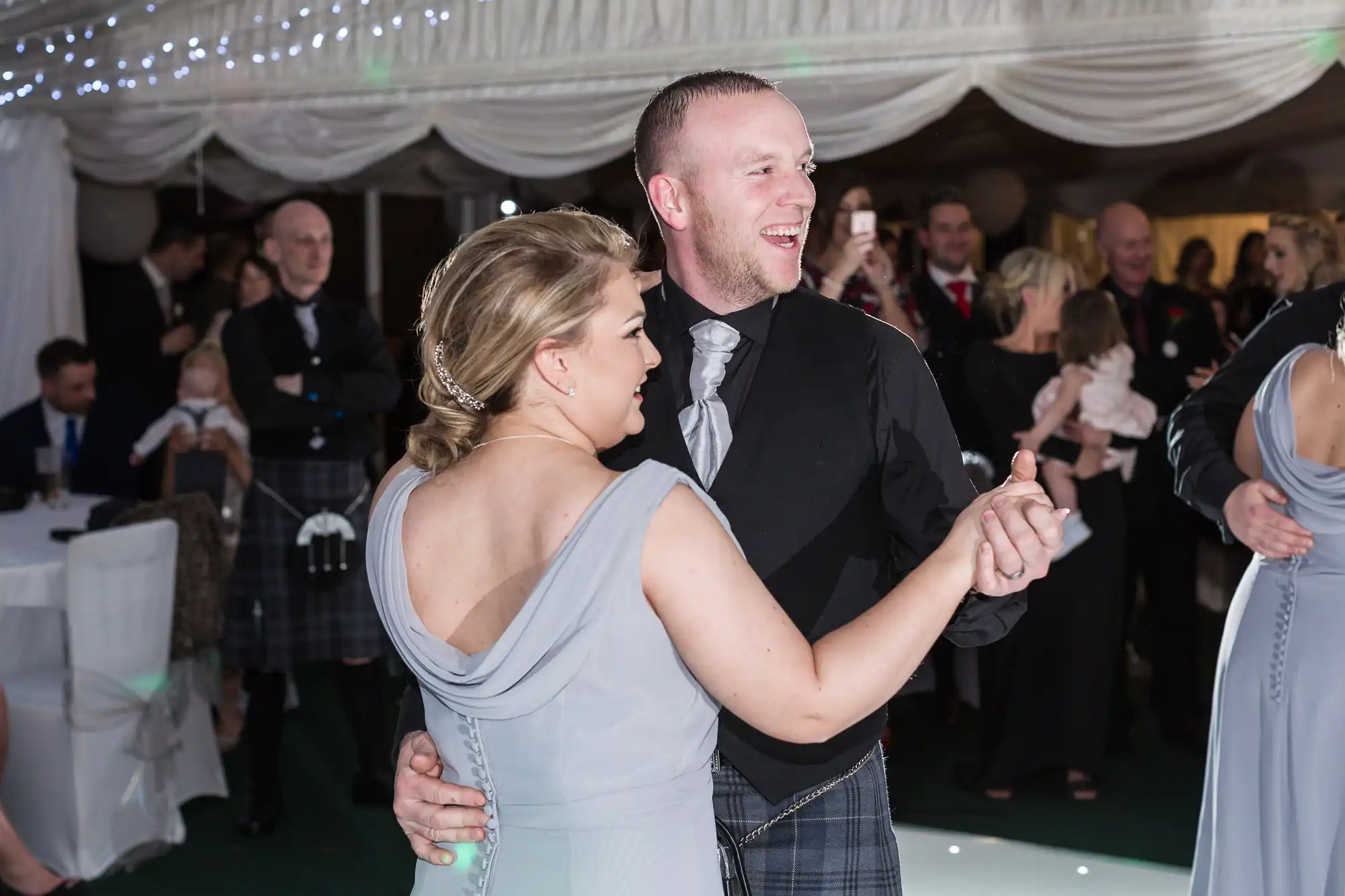 A smiling couple wearing formal attire dances closely at an indoor event. The woman wears a light grey dress with her hair styled up, and the man wears a black jacket, grey waistcoat, and plaid kilt. People in formalwear watch and chat in the background under fairy lights.