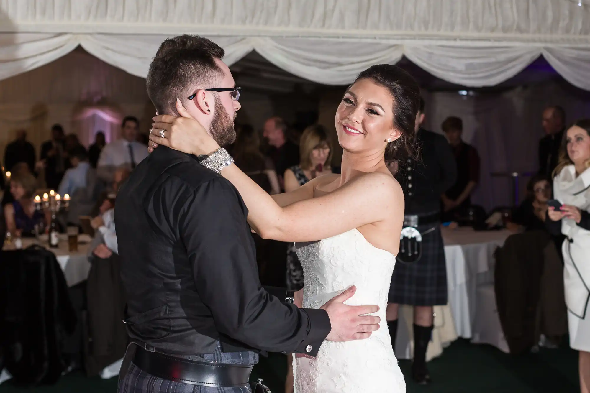 A bride in a white dress smiles while dancing with a groom in black attire at a wedding reception. Guests sit and stand in the background, some watching, under white draped fabric and soft lighting.