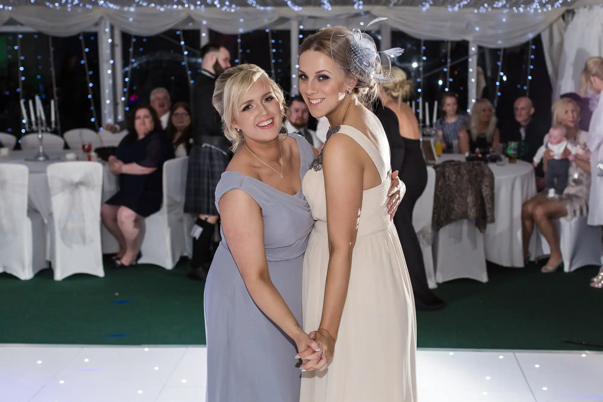 Two smiling women in formal dresses, one in pale grey and one in cream, hold hands and pose for a photo on a white dance floor. Behind them are seated guests, twinkling fairy lights, and white draped decorations at a festive event or wedding reception.