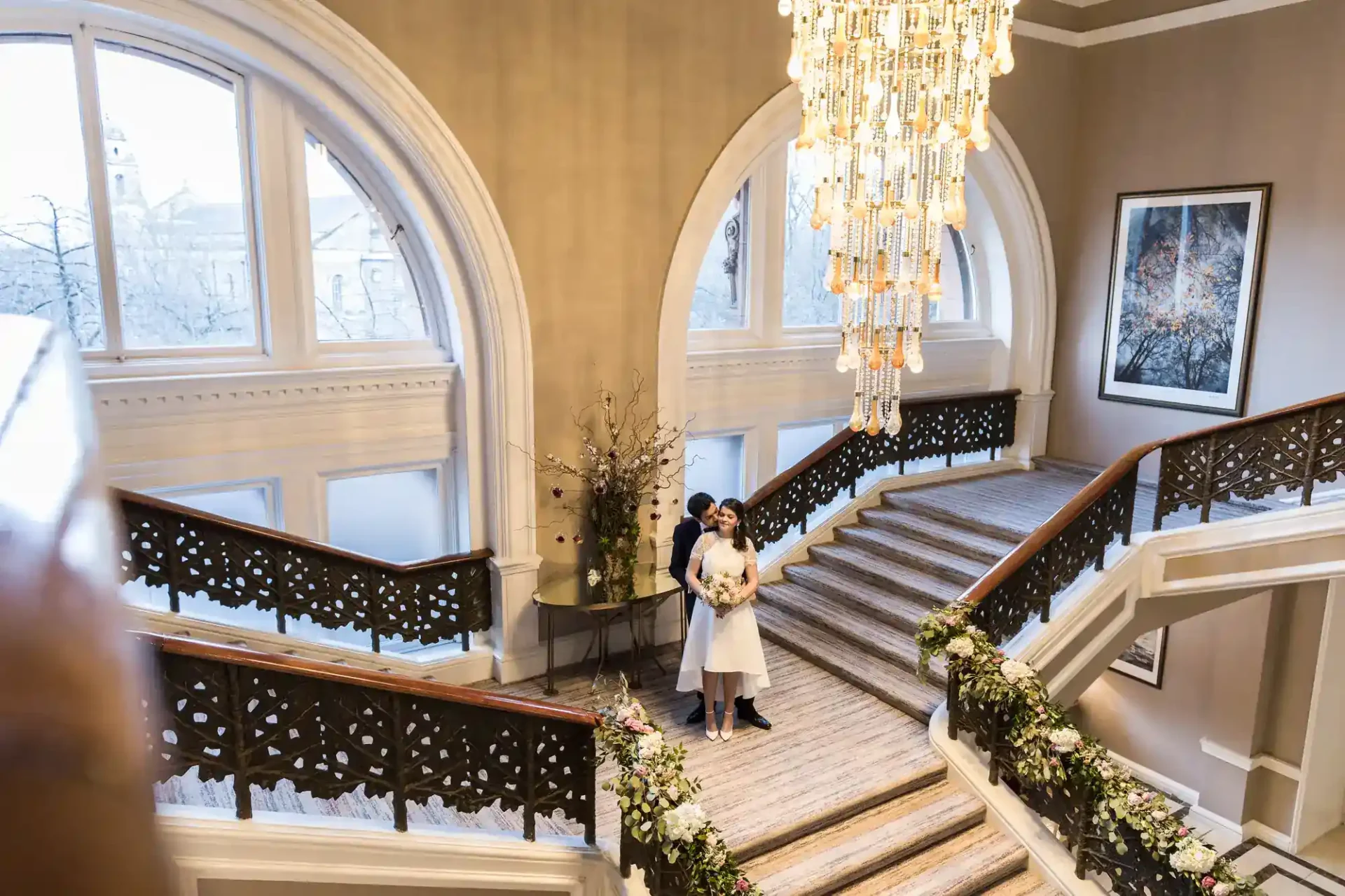 A couple in wedding attire stands on a grand staircase decorated with flowers, beneath a large chandelier and arched windows at the Caledonian Edinburgh.