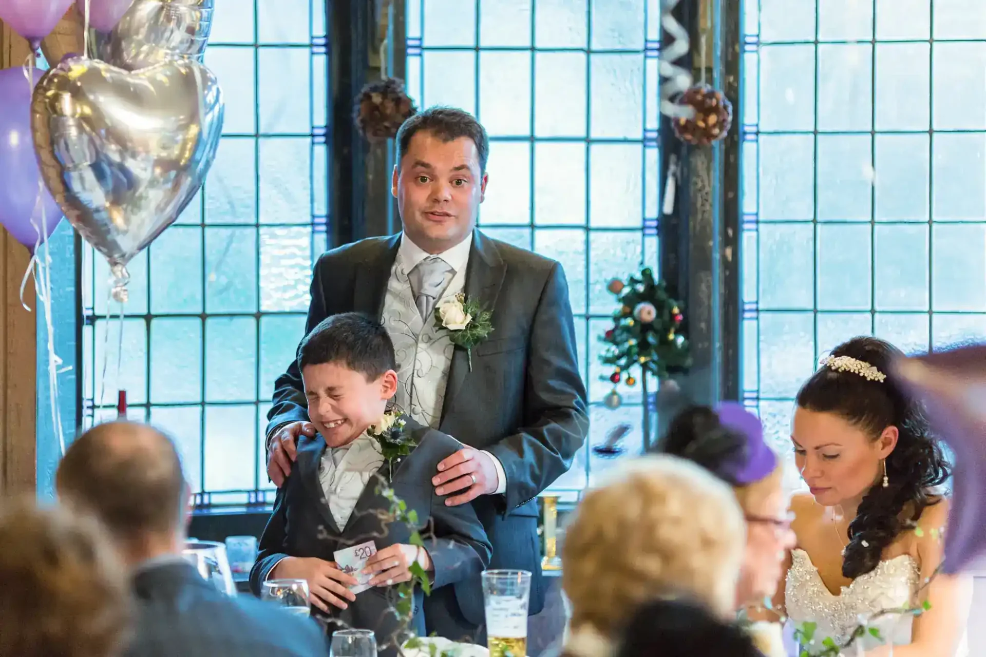 A man in a suit, smiling nervously, stands behind a young boy in formalwear who is laughing with his eyes closed. They are at a wedding reception at The Lodge at Carfraemill, with guests seated, a bride in a white dress to the right, and heart-shaped balloons and decorations visible.