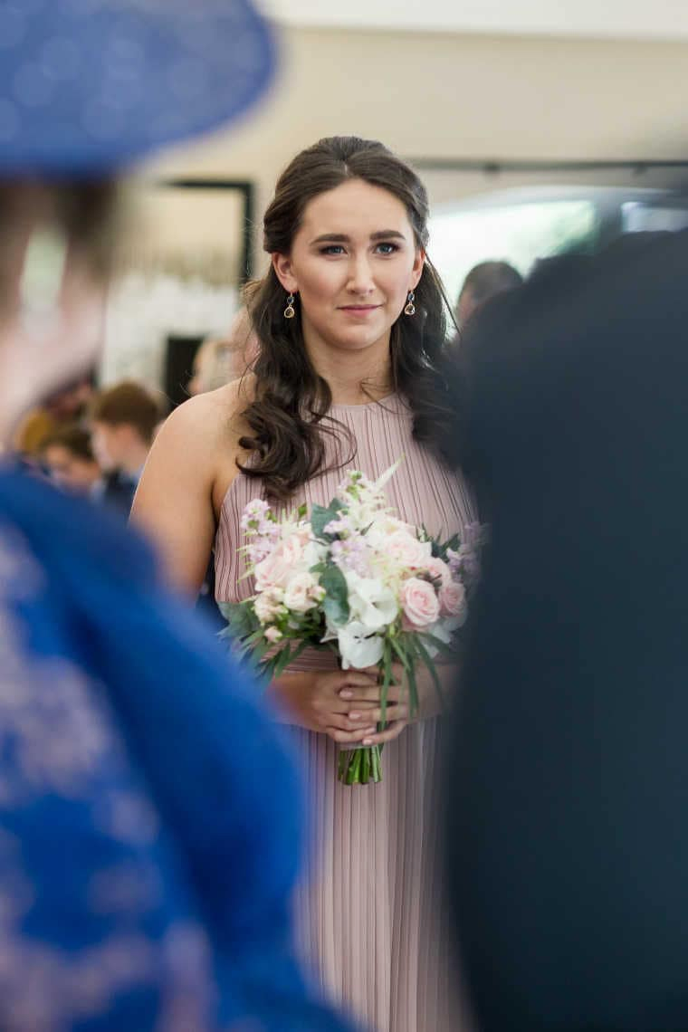 Young woman in a pleated pink dress holding a bouquet of flowers, looking thoughtful at a social event.