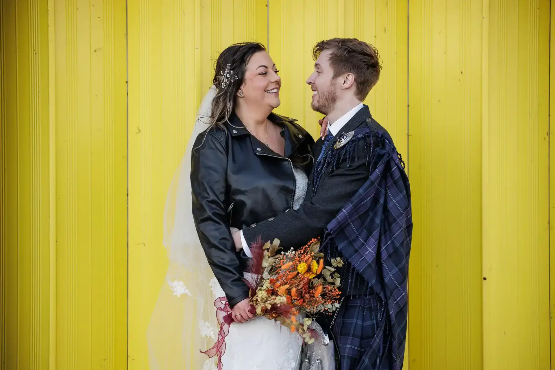 A bride in a white dress, black leather jacket, and veil holds a bouquet of autumn flowers. She smiles at a groom in a kilted suit and checked shawl. They stand facing each other, lovingly gazing, against a bright yellow wooden wall.
