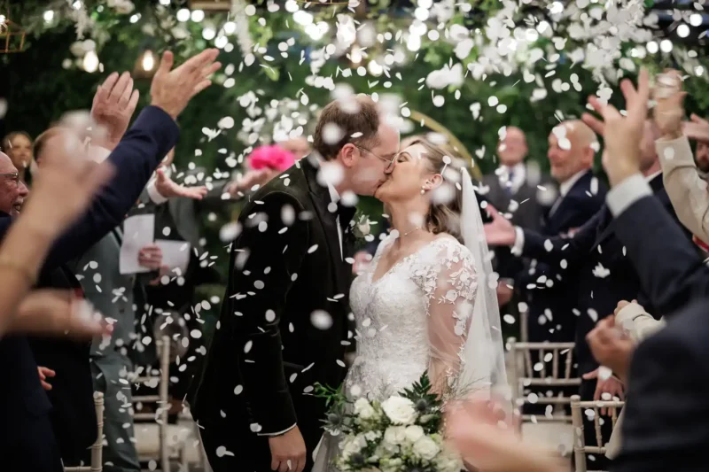 A bride and groom kiss at their wedding ceremony as guests throw white confetti around them.