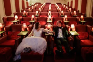 A bride and groom in wedding attire sit holding hands in red velvet seats of an empty theater, surrounded by small lit lamps on tables.