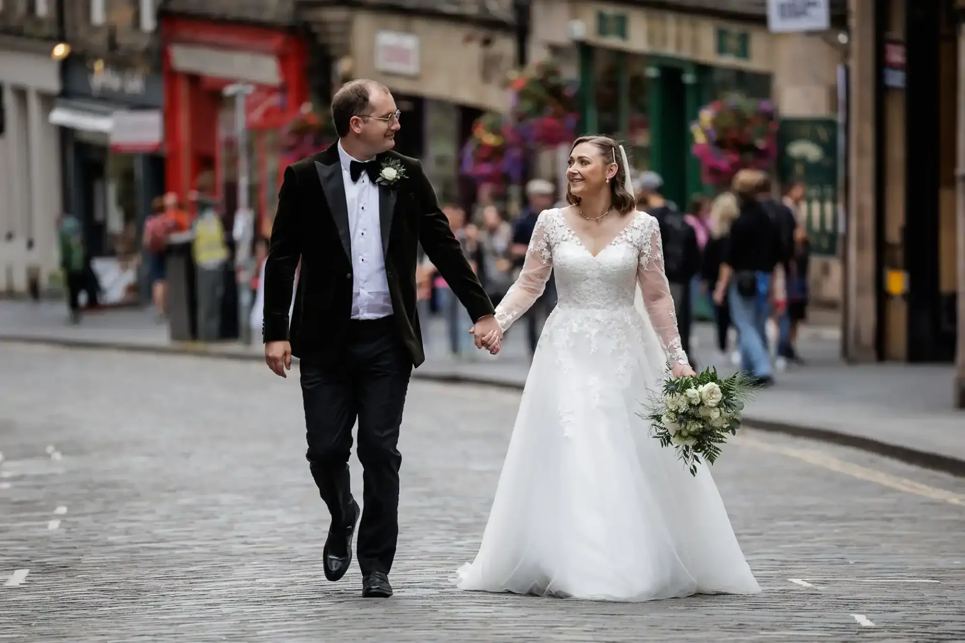 A bride and groom in formal wedding attire walk hand in hand down Cockburn Street, smiling at each other, with people and shops in the background.