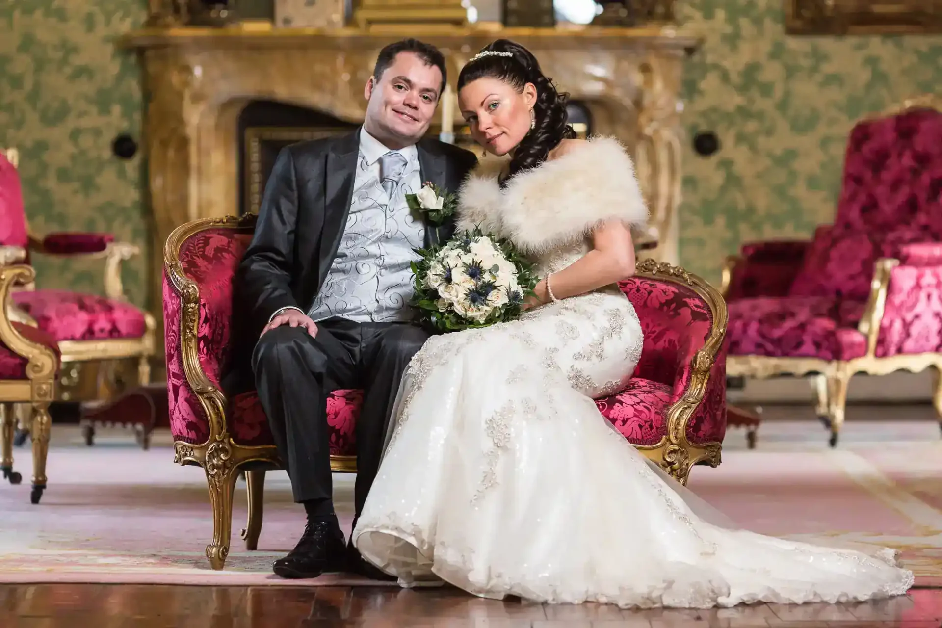 A bride in a white gown and faux fur wrap sits closely with a groom in a dark suit on an ornate red and gold sofa. She holds a white bouquet; they both smile. The elegant room at Thirlestane Castle has rich décor, patterned wallpaper, and a marble fireplace in the background.