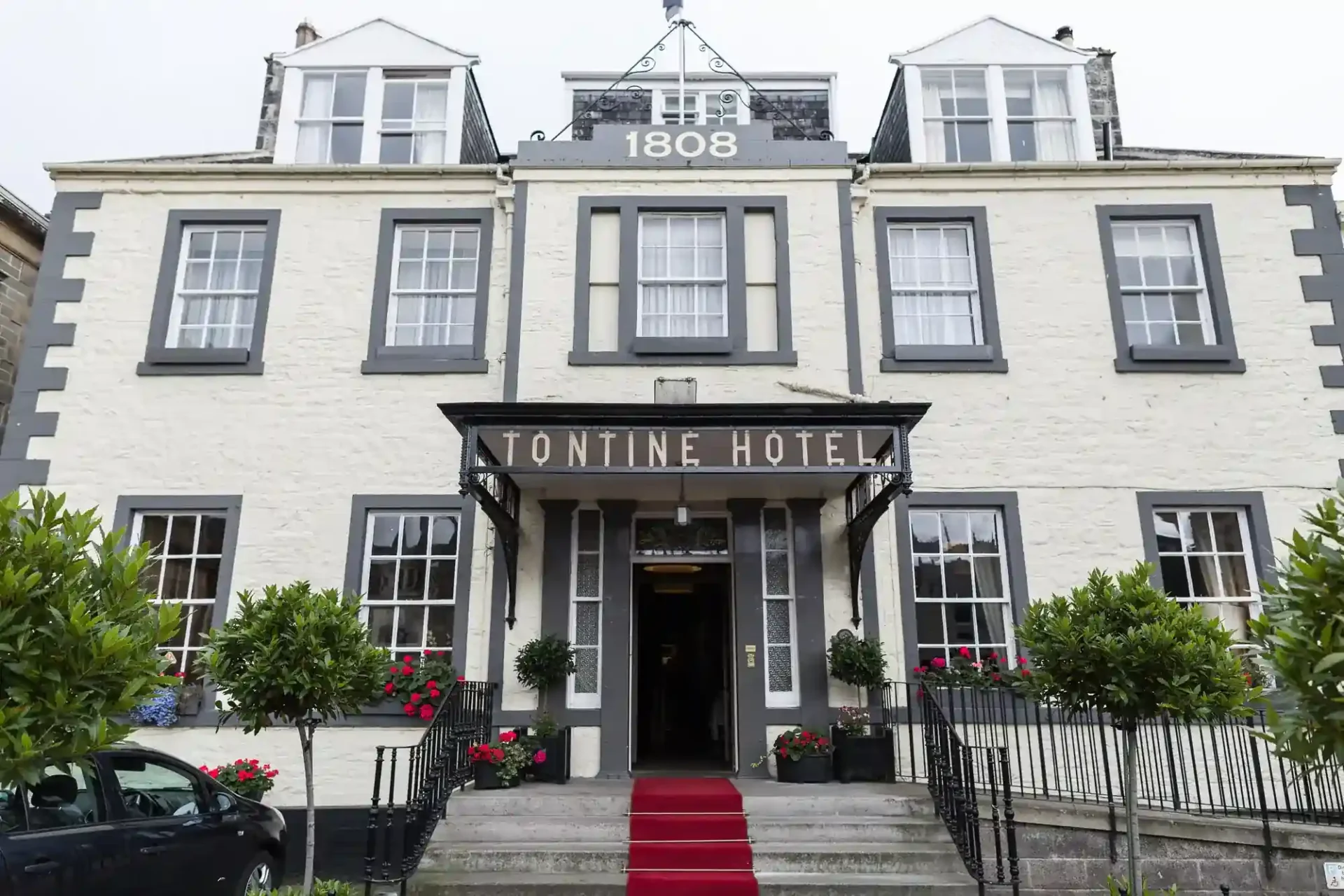 The Tontine Hotel, a cream-painted, historic hotel with grey trim, reading “Tontine Hotel” above the entrance. The building has large windows, a red carpeted front stairway, greenery in planters, and “1808” displayed at the roof peak, indicating its establishment year.