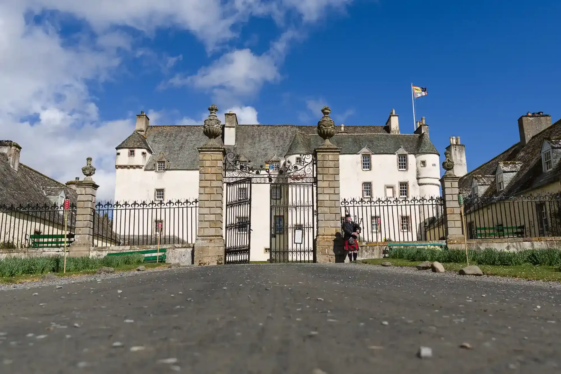 A wide stone path leads to a grand white historic building, Traquair House, with a tiled roof, stone pillars, and ornate iron gates. Two people stand near the entrance. The sky is blue with scattered clouds and a flag flies atop the building.