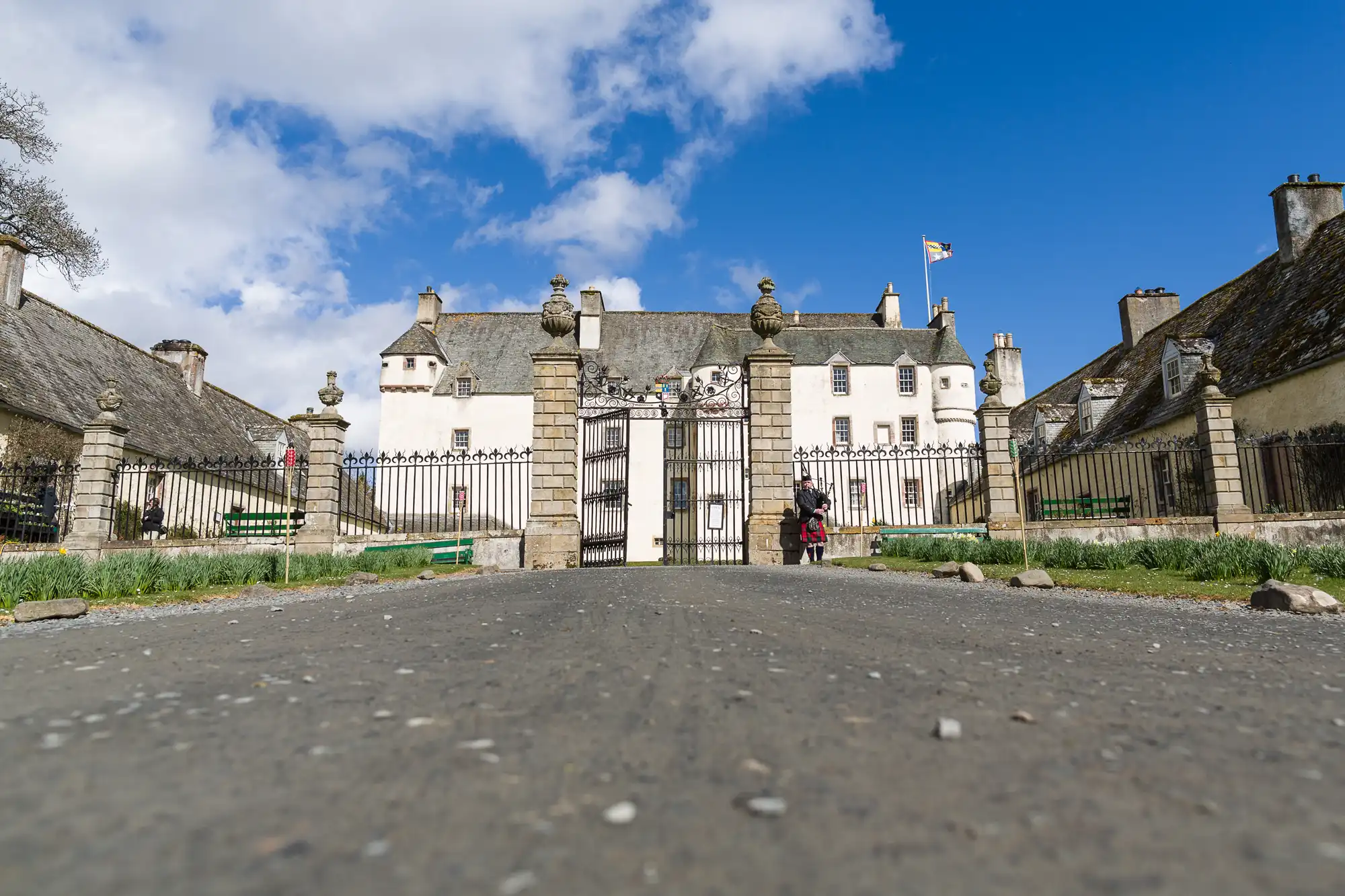 View of a grand, white historic manor with a Scottish flag, seen from a low angle. Stone pillars and a black iron gate enclose the entrance. Two people stand at the gated entrance, with gardens and blue sky in the background, by award-winning Traquair House wedding photographer.