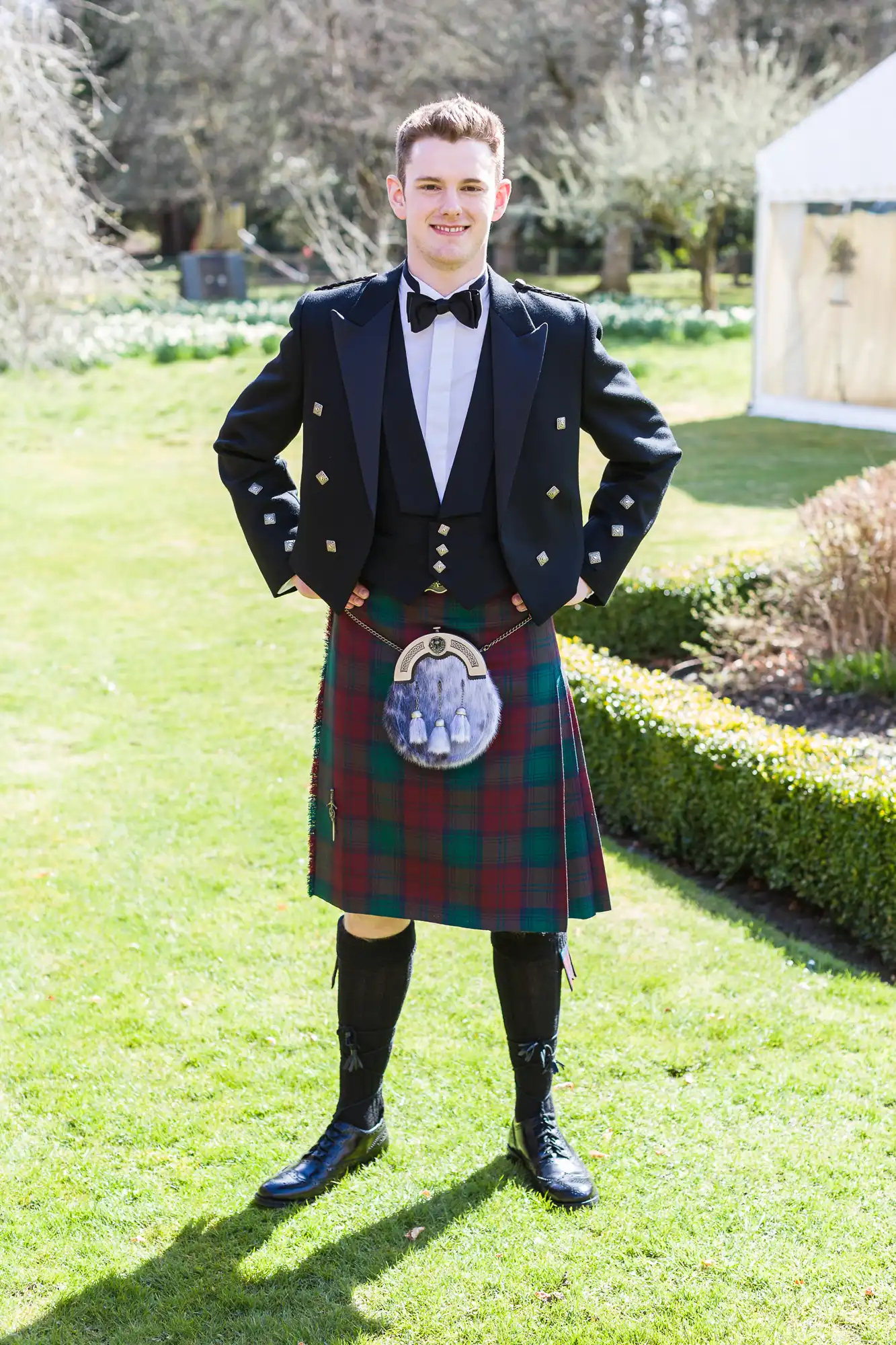 A young man stands outdoors on a lawn, wearing a traditional Scottish outfit—black jacket, white shirt, bow tie, red and green tartan kilt, sporran with a photo, long socks, and black shoes. There’s a garden and marquees in the background.