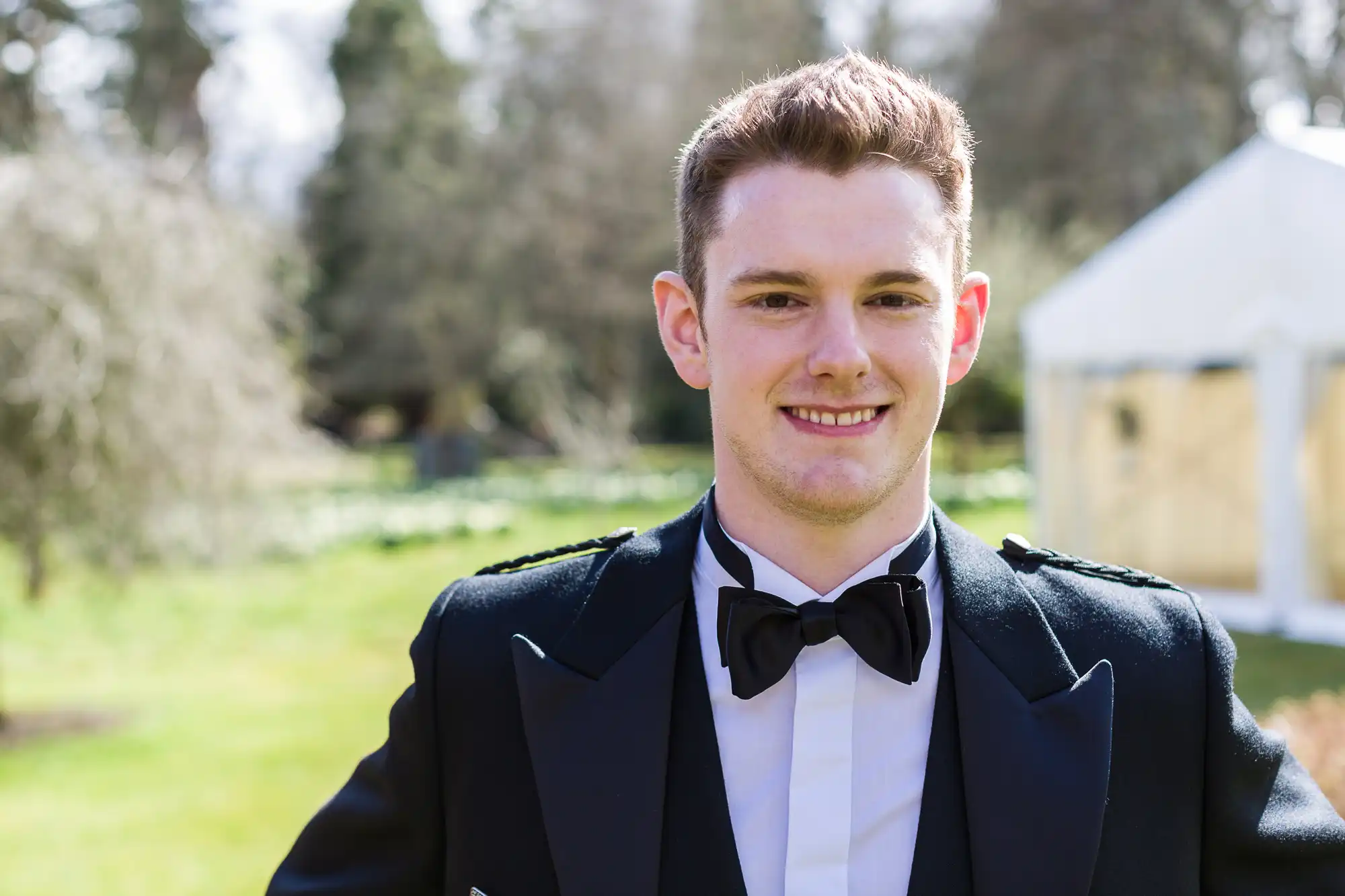 A young man with short brown hair is smiling outdoors. He is wearing formal attire: a black bow tie, white shirt, and a traditional black jacket. The background shows green grass, trees, and a white event marquee in soft focus.