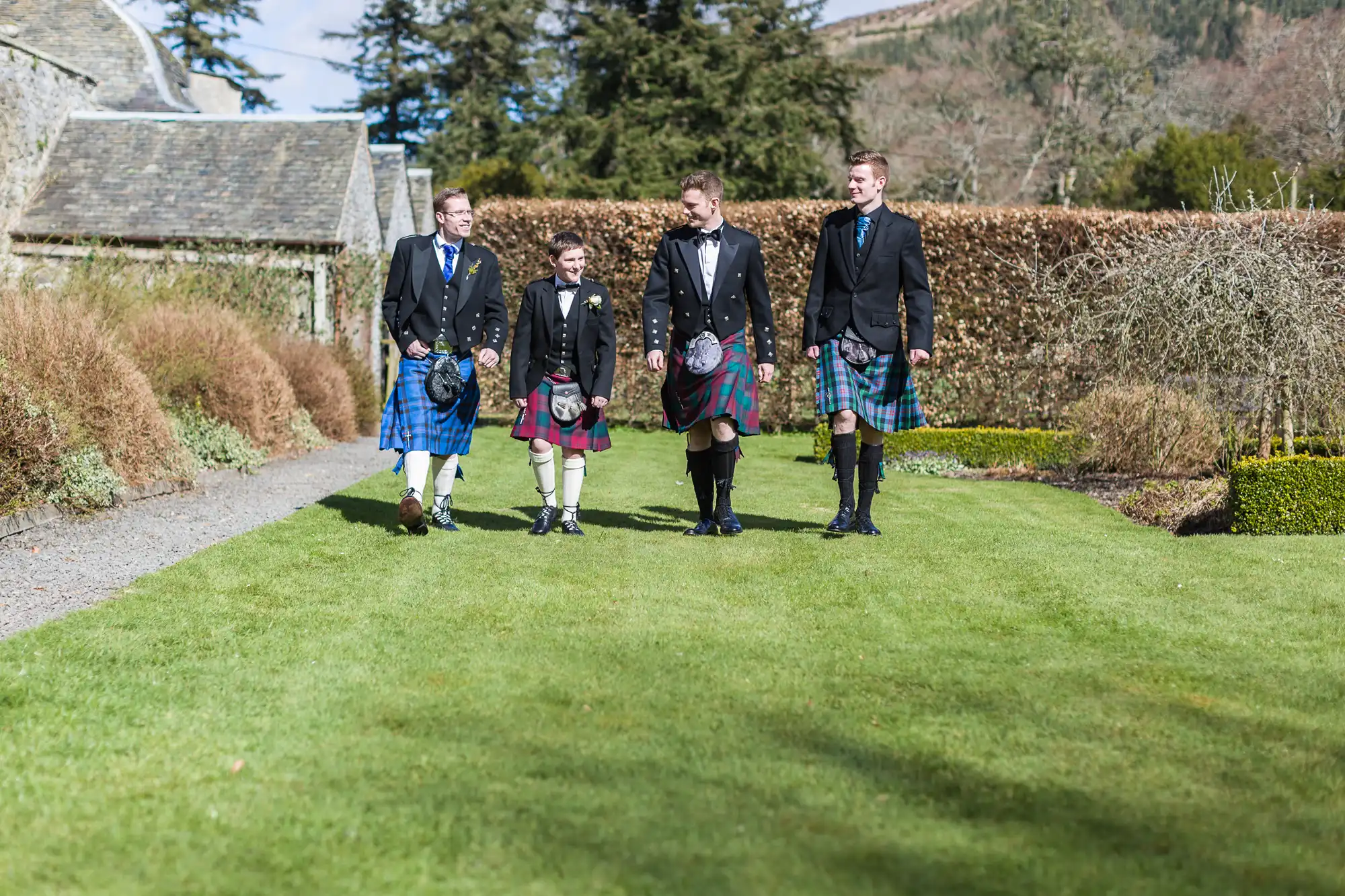 Four men walk outside on green grass, all wearing traditional Scottish kilts, jackets, and sporrans. They are smiling and talking. Behind them are trimmed hedges, trees, and a stone building under a partly cloudy sky in a rural setting.