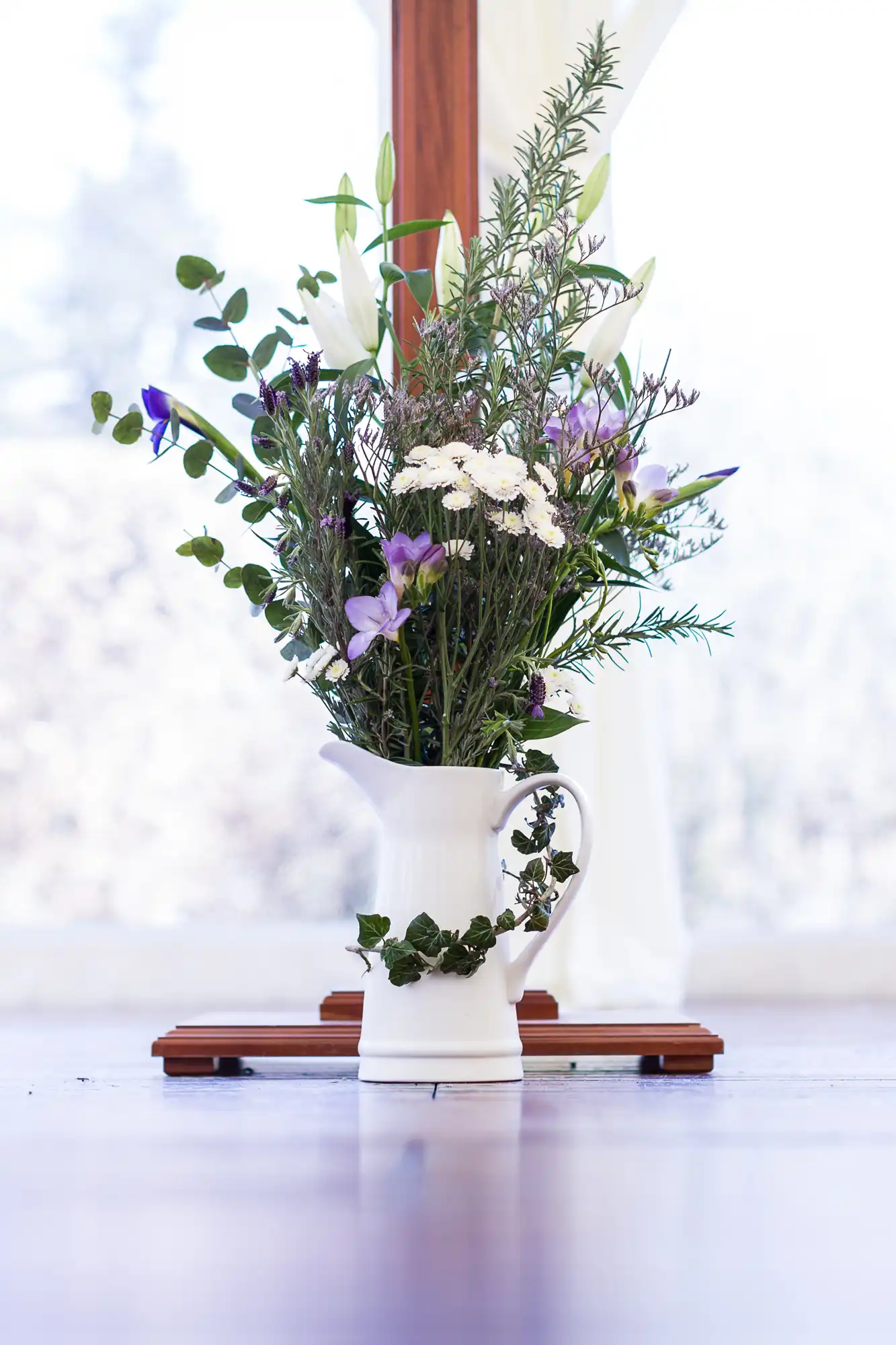 A white ceramic jug filled with a bouquet of assorted flowers and greenery, including white lilies, purple blooms, and leafy stems. The jug sits on a smooth surface in front of a bright window with wooden frames and sheer curtains.