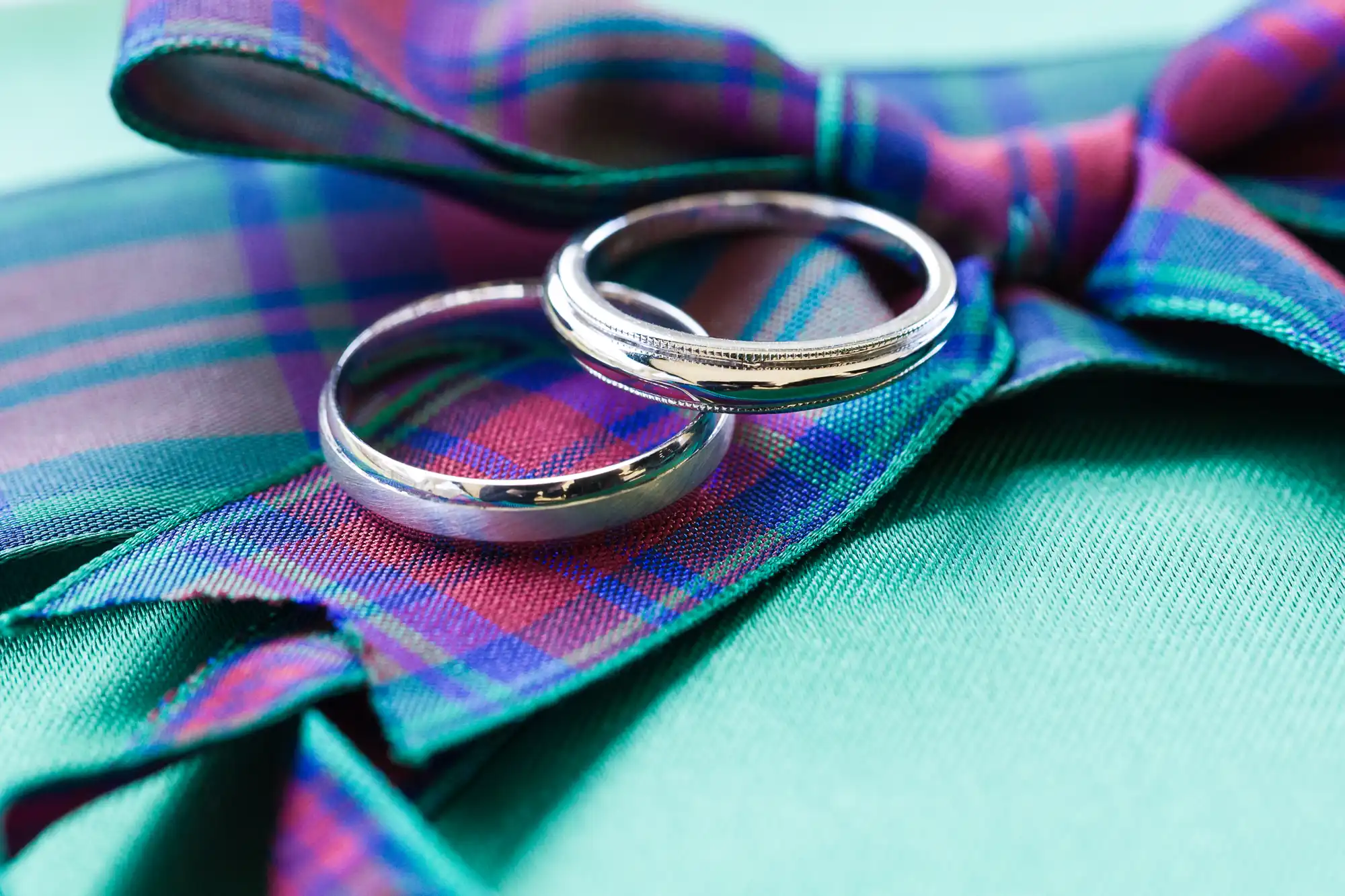 Two silver wedding rings rest on a purple, blue, and green tartan-patterned bow tie set against a teal fabric background. The rings are shiny, simple in design, and placed to intersect, symbolising unity or partnership.