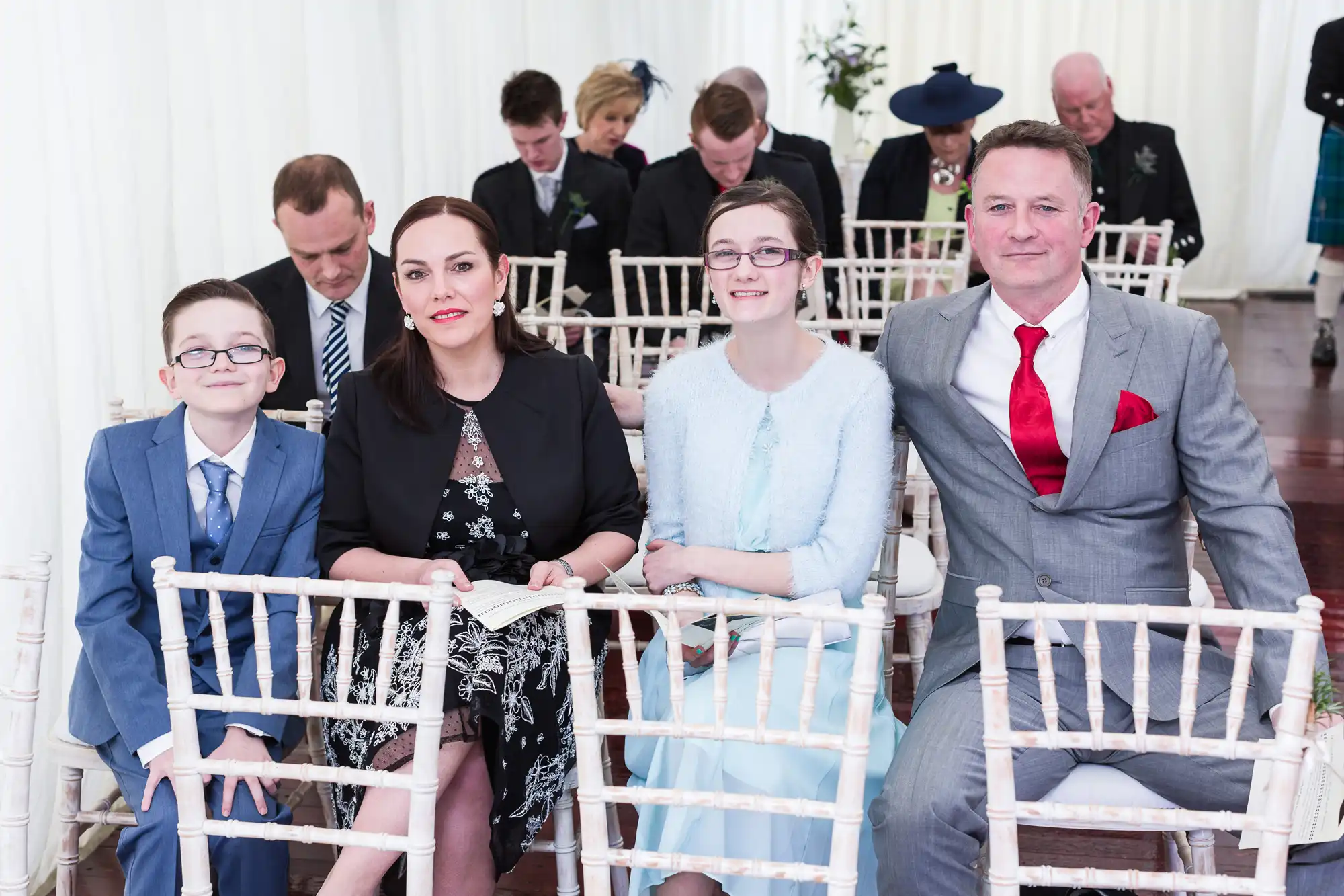 Four people sit in the front row of white chairs at an event, dressed formally; a smiling boy in glasses, a woman in black, a girl in light blue, and a man in a grey suit with a red tie. Other guests sit behind them, some looking down.