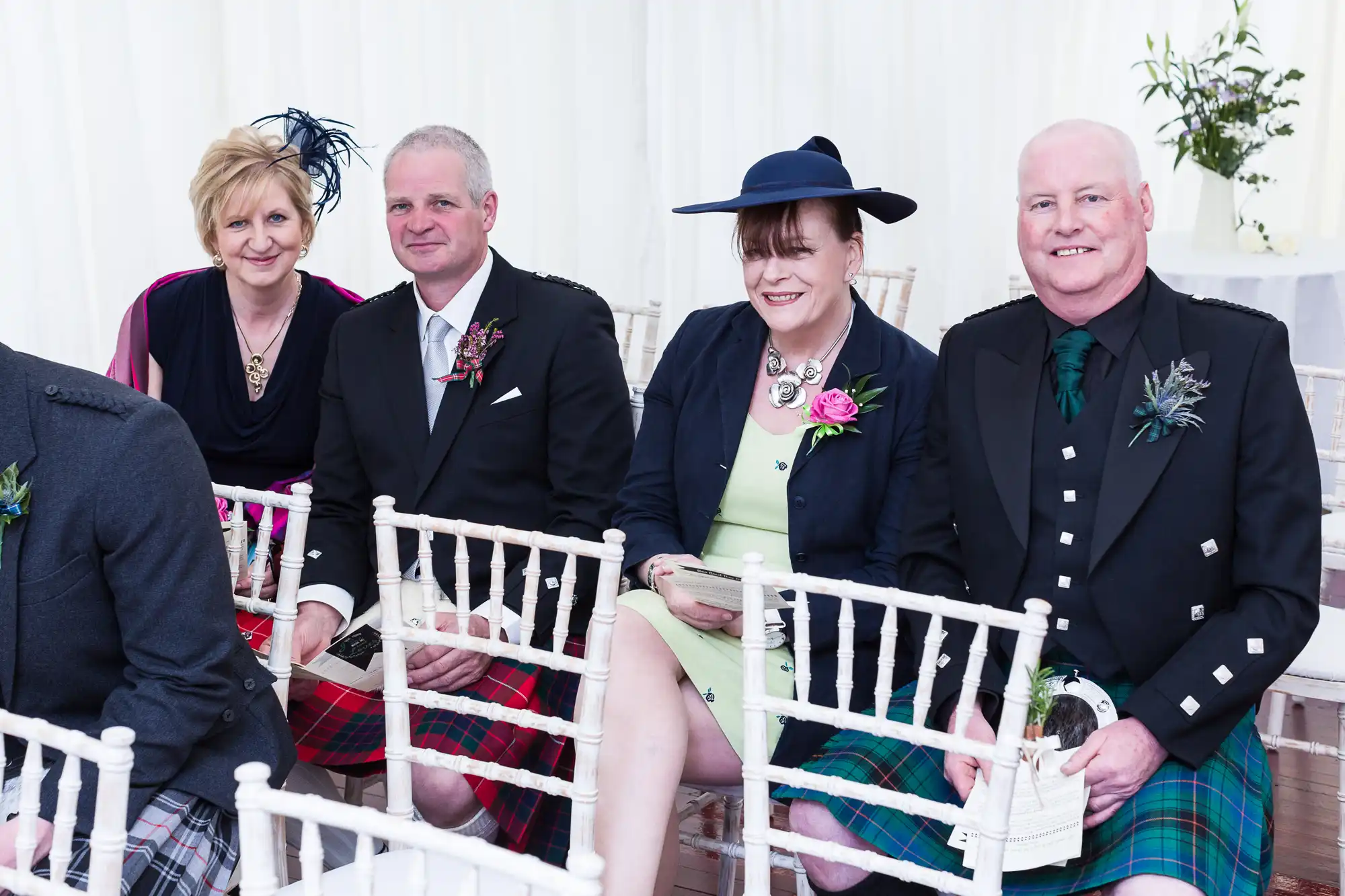 Four people sit indoors on white chairs at a formal event. The two men wear kilts and dark jackets; the women wear dresses with jackets and hats or fascinators. All are smiling and holding papers, with wedding-style decorations in the background.