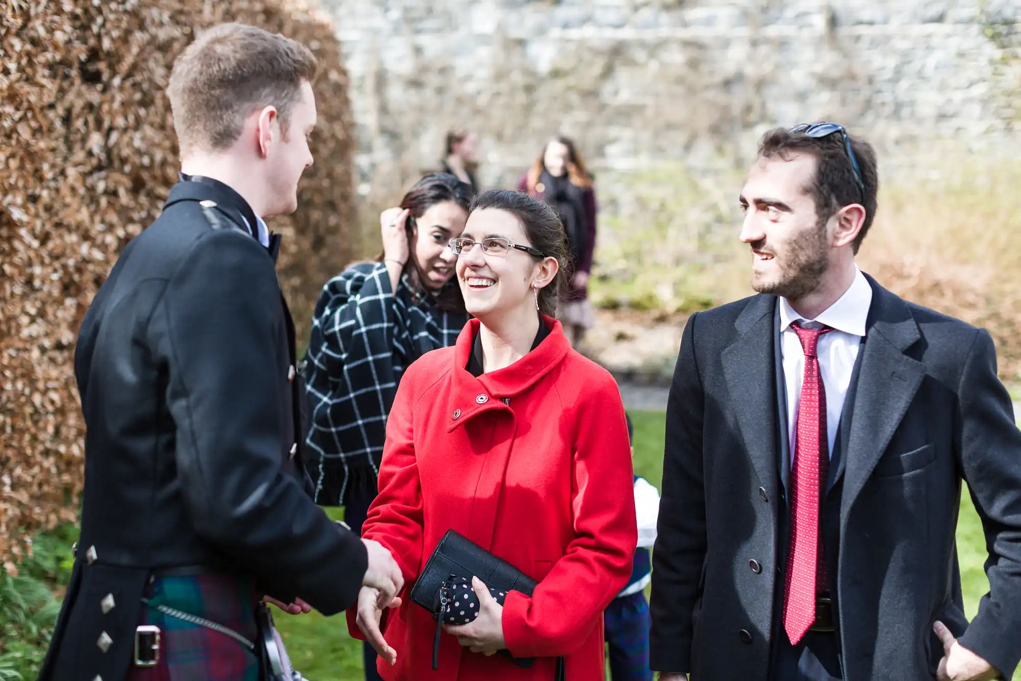 A man in a kilt shakes hands with a smiling woman in a red coat, holding a clutch bag. Another man in a dark coat and red tie stands beside her, smiling. People and greenery are in the background, with a stone wall behind them on a sunny day.