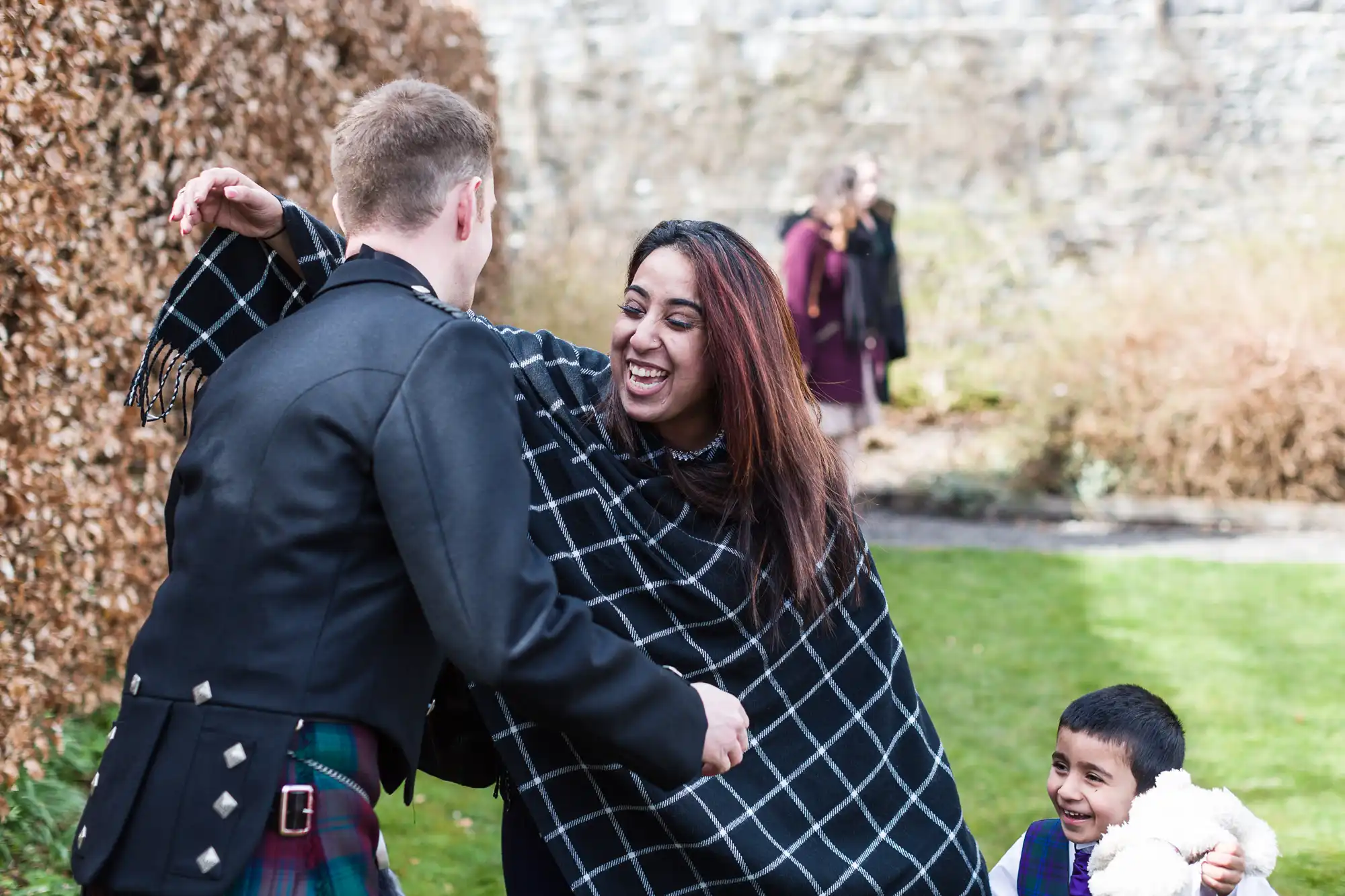 A smiling woman in a black-and-white chequered shawl hugs a man in a traditional Scottish kilt outfit outdoors. A young boy holding a soft toy looks up and smiles nearby. Blurred plants and people are in the background.