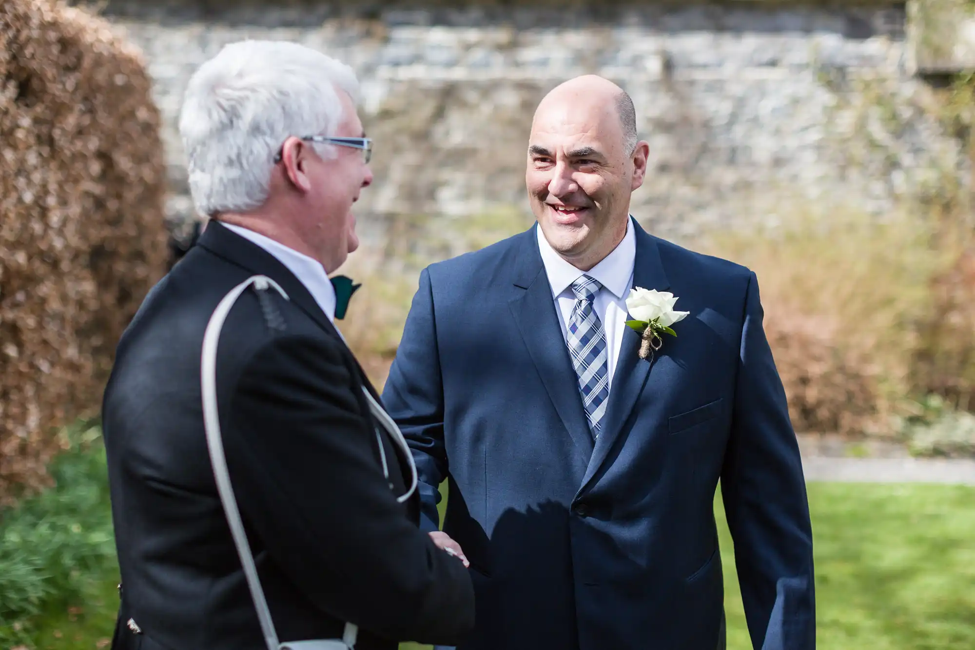 Two men in formal attire shake hands outdoors. One wears a navy suit with a striped tie and a white rose buttonhole, smiling warmly. The other, with grey hair and glasses, is dressed in a black jacket with white trim. Sunlit garden and stone wall in background.