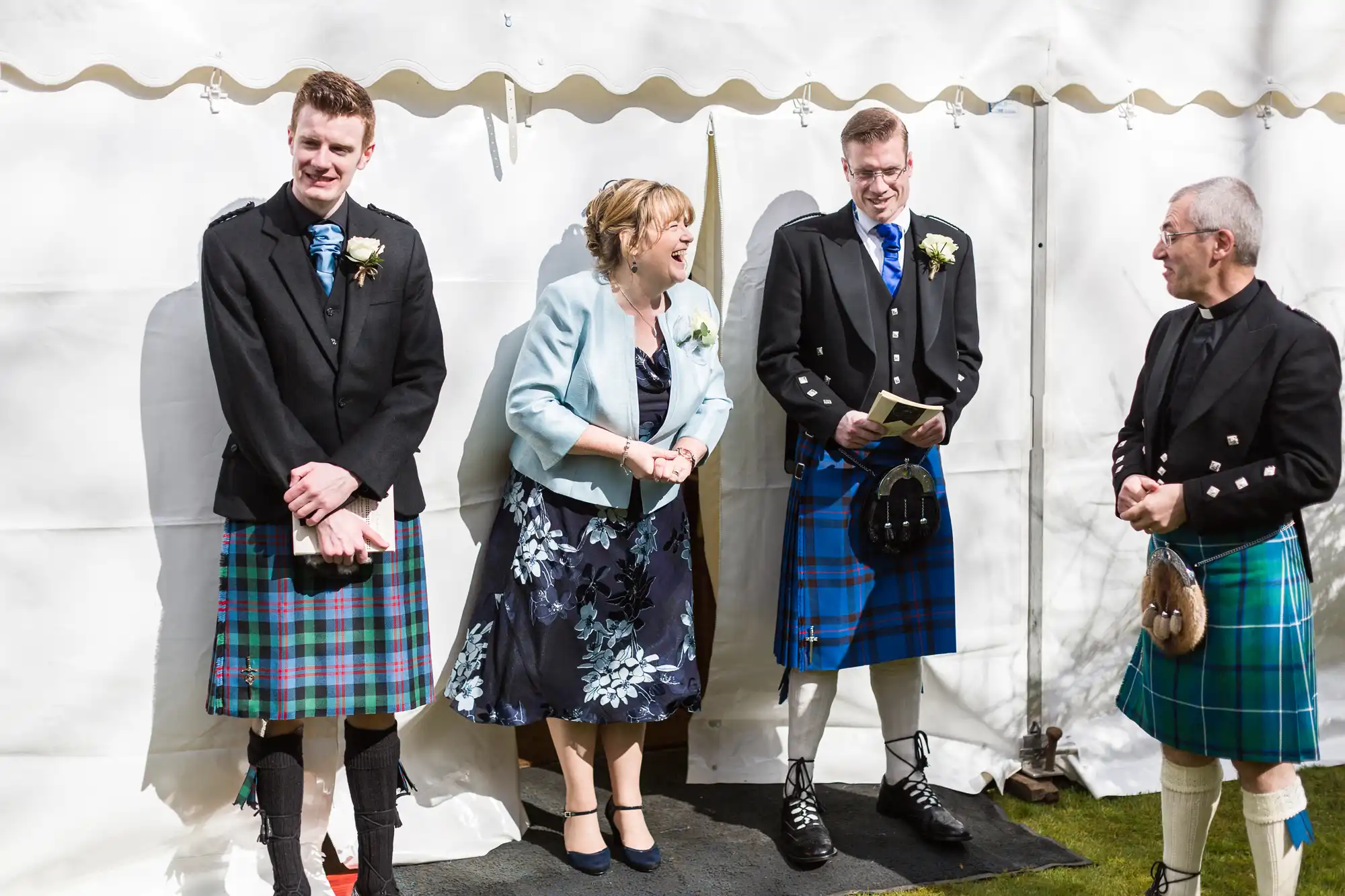 Four adults stand and chat outside under a white marquee. Three men wear black jackets with tartan kilts in blue and green patterns; one woman wears a pale blue jacket and floral dress. They all smile and appear to be enjoying a light-hearted moment together.