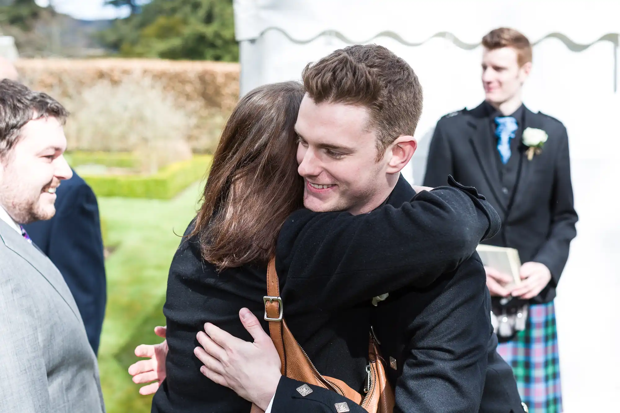 A young man in a black jacket smiles as he hugs a woman outdoors at a formal event. Two men in kilts and suits stand nearby, one holding a book. The group is in a garden with trimmed hedges and a white marquee in the background.