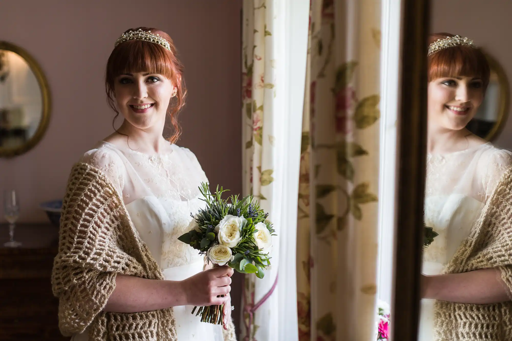 A smiling bride with red hair, wearing a tiara, white dress, and beige shawl, holds a bouquet of white and purple flowers whilst standing beside a window with floral curtains. Her reflection is visible in the glass.