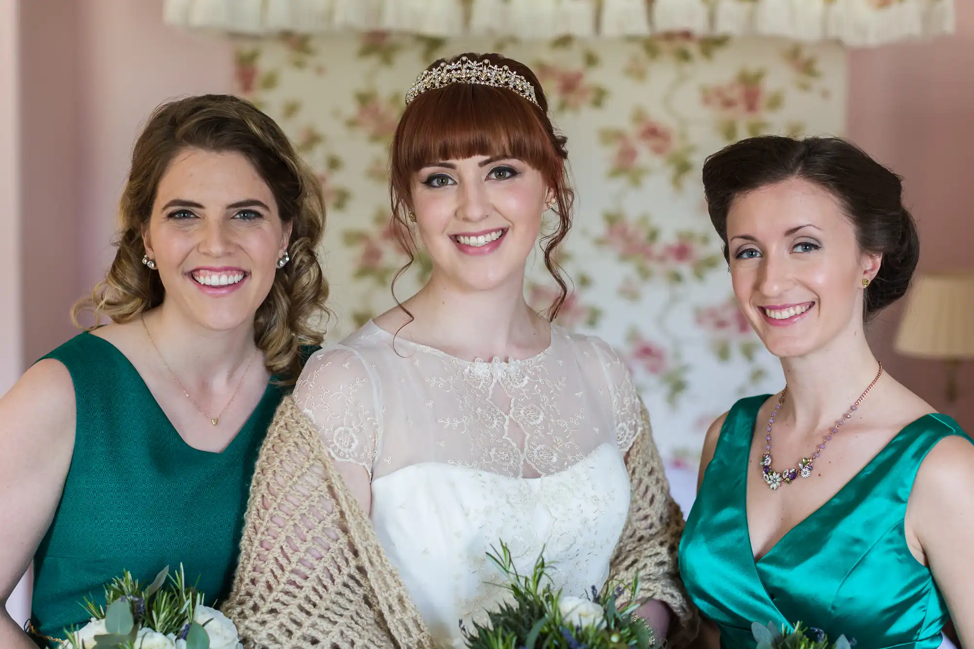 Three women pose indoors, smiling. The centre woman wears a white wedding dress, tiara, and a lace shawl, holding a bouquet. The two women beside her wear teal dresses and also hold bouquets. The background features floral wallpaper and soft lighting.