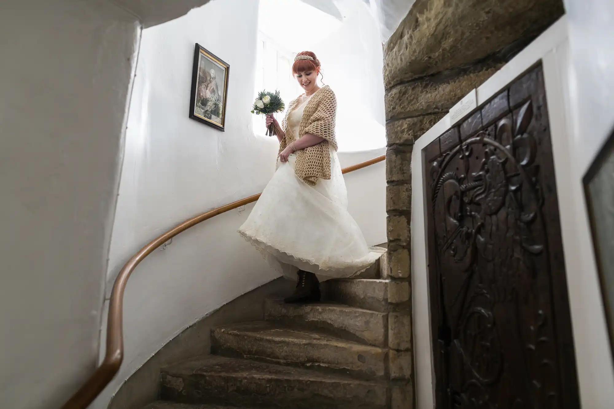 A bride in a white dress and beige shawl holds a bouquet whilst smiling, descending a narrow, curved stone staircase with white walls, a framed picture, and a wooden door with ornate carvings. Natural light shines from above.