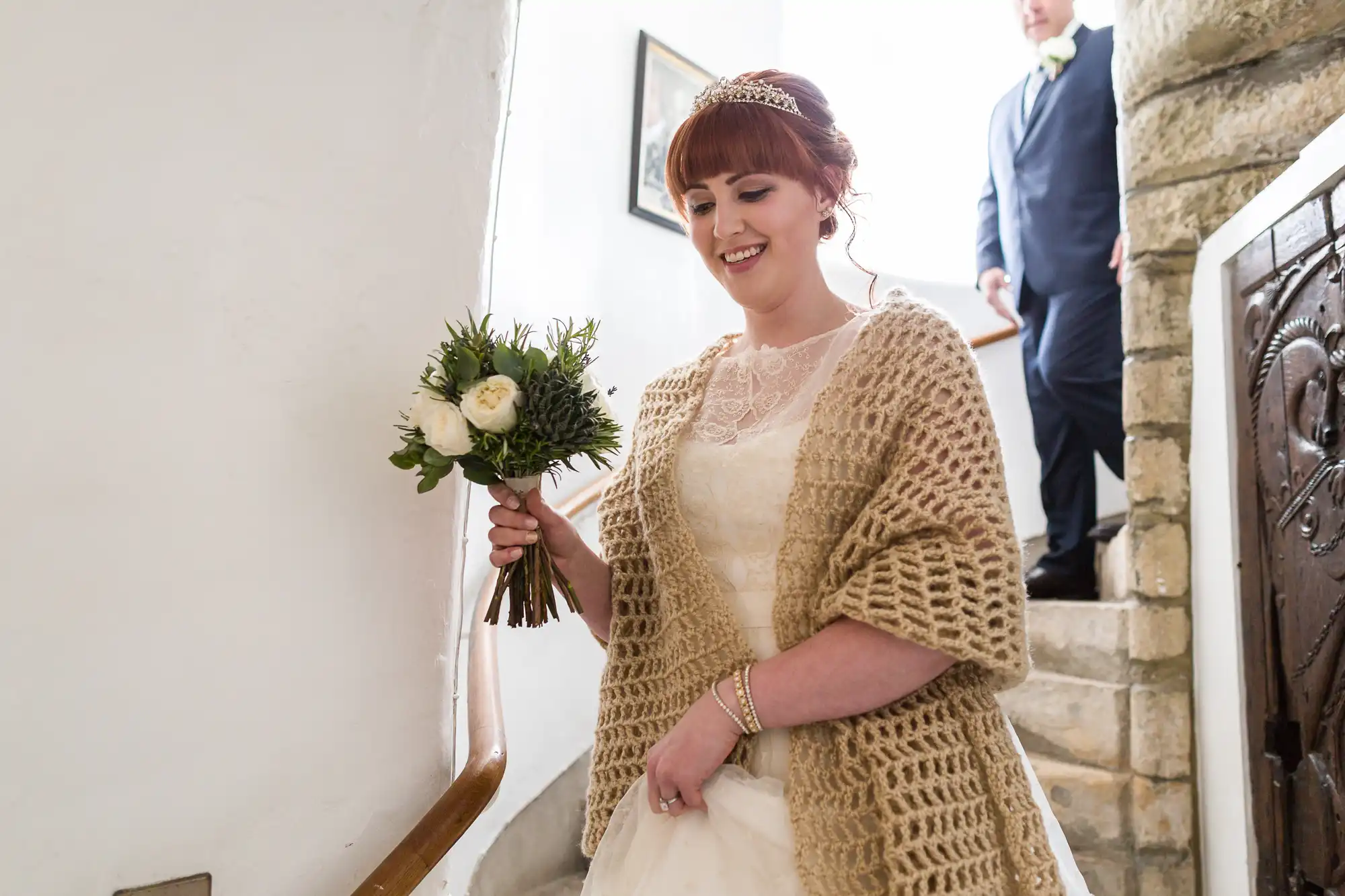 A bride with red hair, wearing a cream shawl over her lace wedding dress, holds a bouquet of white flowers and greenery whilst smiling and walking up white stairs. A man in a suit is descending the stone steps behind her.