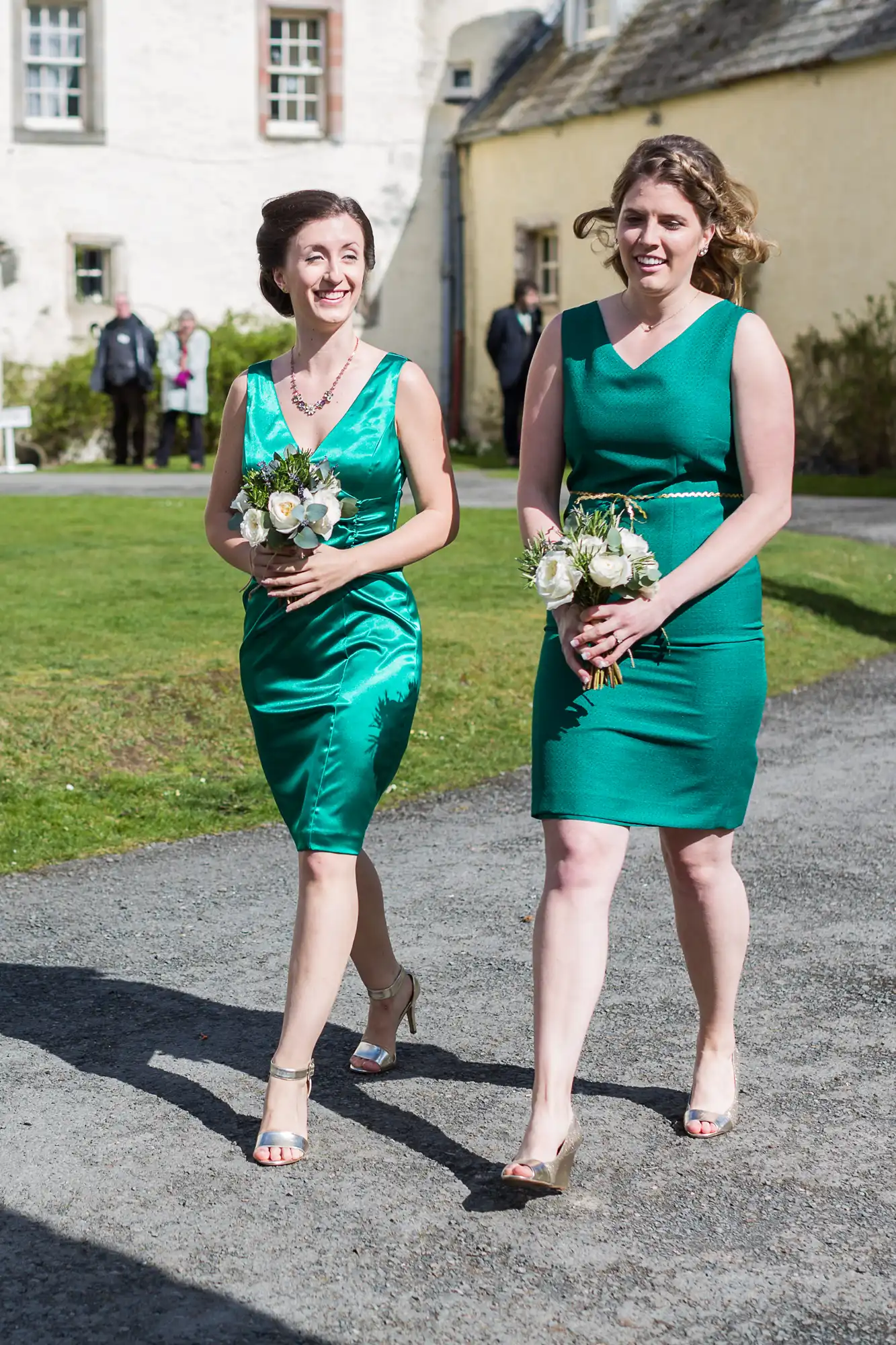 Two women in sleeveless green dresses walk side by side outdoors on a sunny day, holding small posies of white flowers. They are smiling, wearing sandals, and appear to be part of a wedding party. A stone building and lawn are in the background.