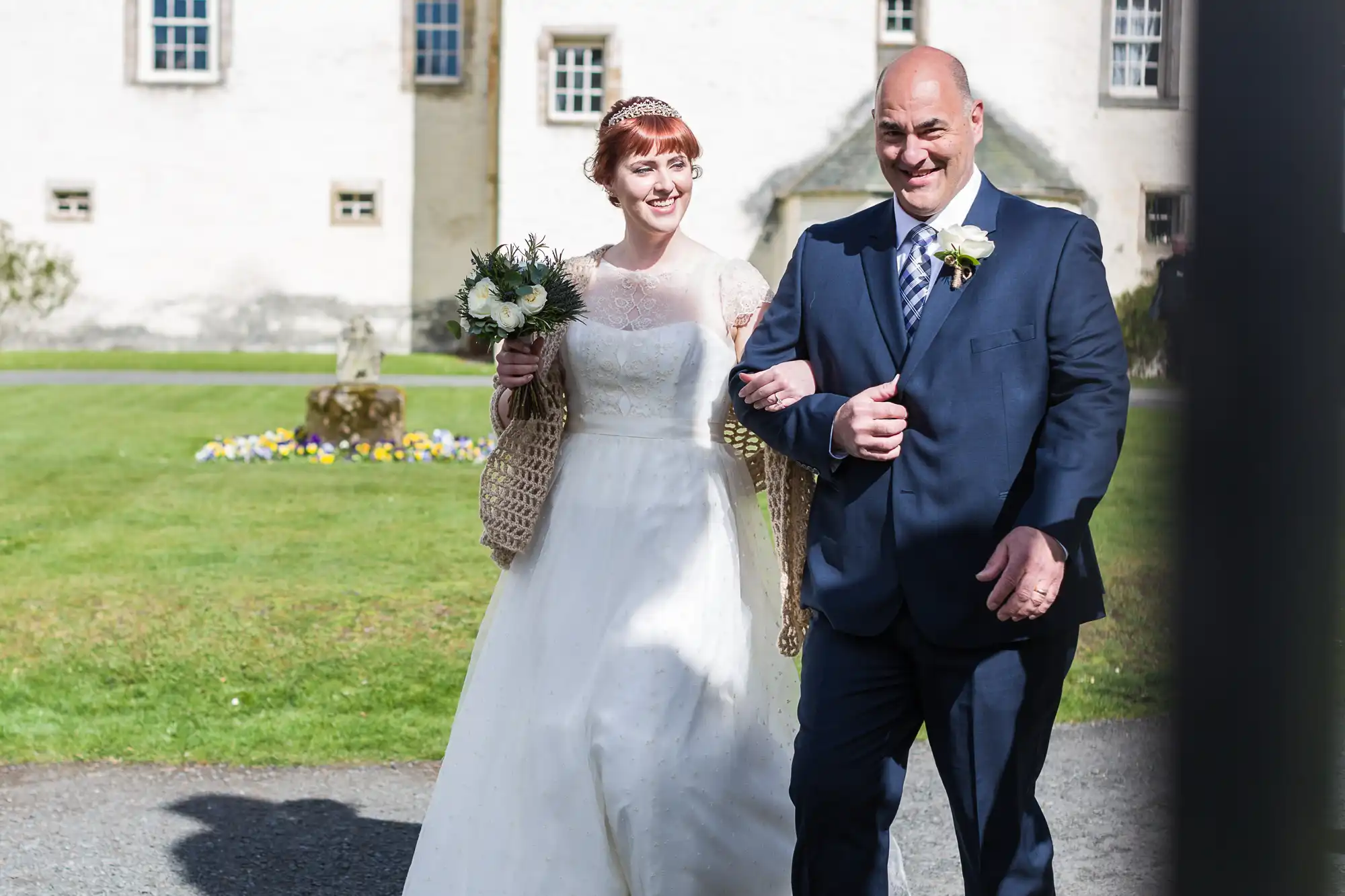 A bride in a white gown with lace sleeves and a tiara walks arm-in-arm outdoors with a man in a navy suit. Both are smiling. The bride holds a bouquet; they are in front of a white building with a green lawn and flowerbed in the background.