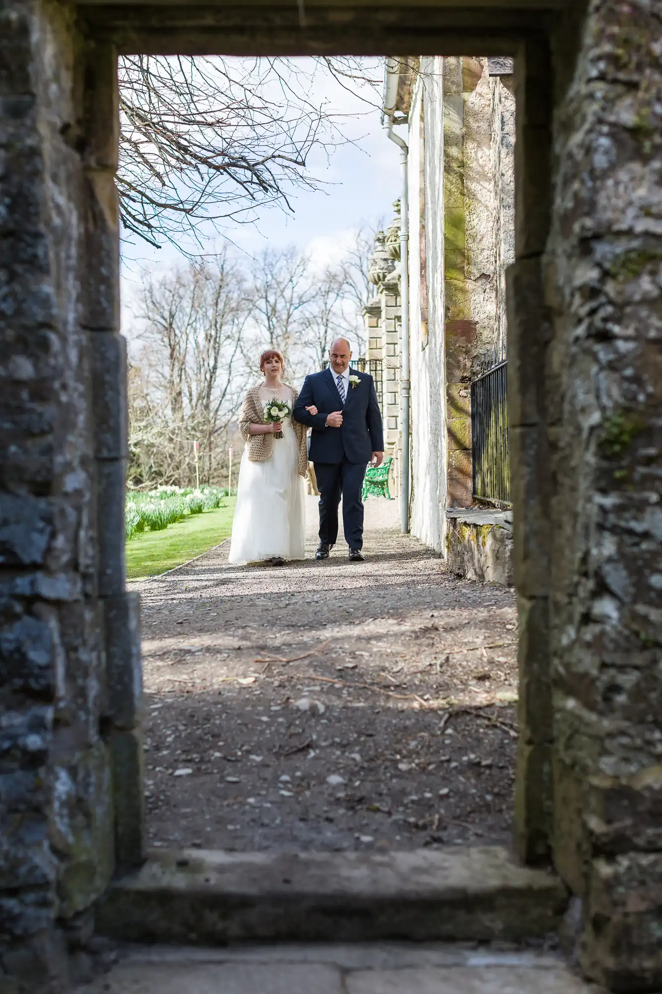 A bride in a white dress and tan shawl walks arm-in-arm with an older man in a suit along a stone path outside, seen through a stone archway. Bare trees and stone walls are in the background under a clear sky.