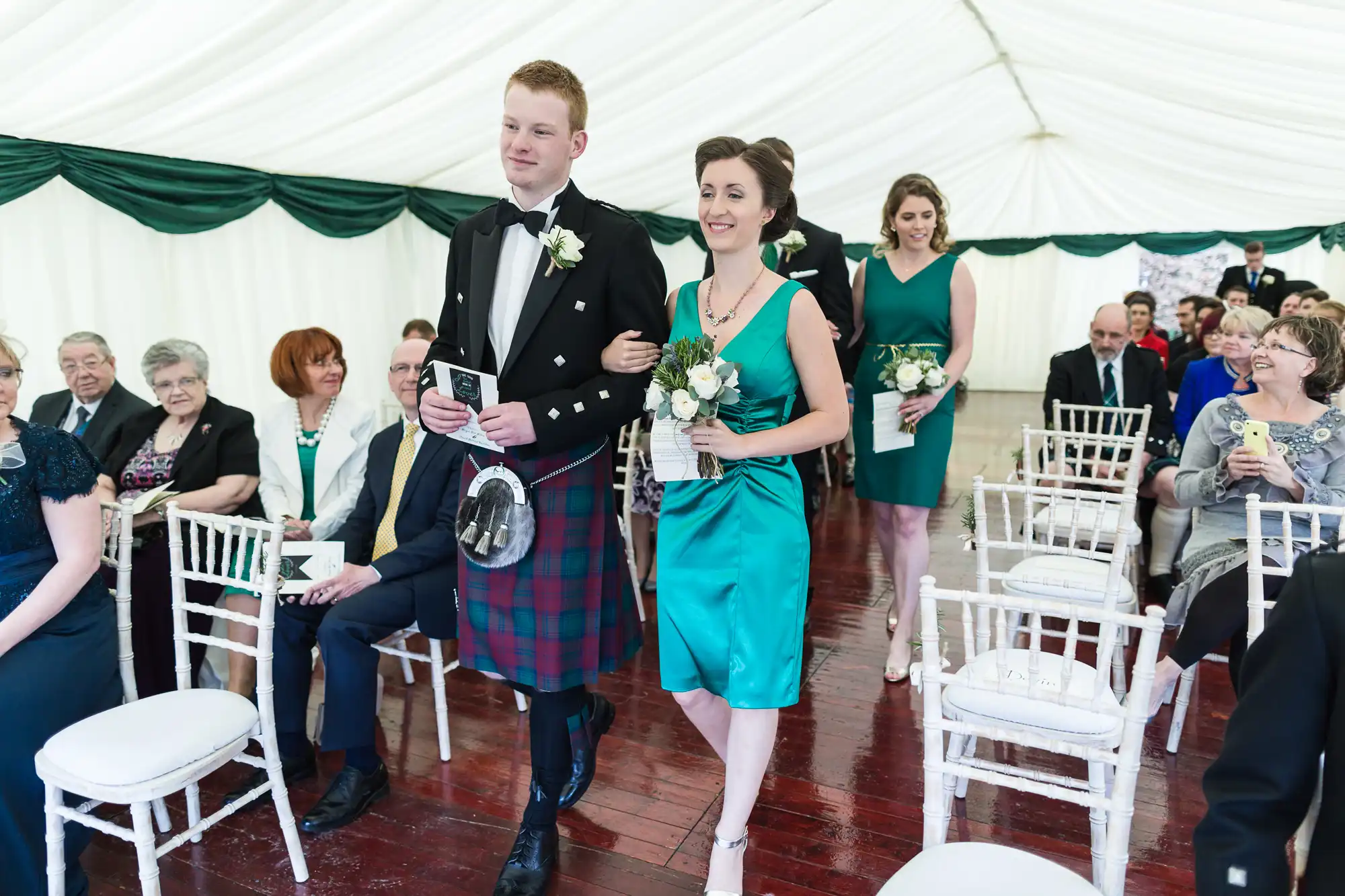 A man in a kilt and black jacket walks arm-in-arm with a woman in a teal dress holding flowers down the aisle of a white marquee, followed by another bridesmaid. Seated guests watch and smile, some holding programmes, in a celebratory wedding setting.