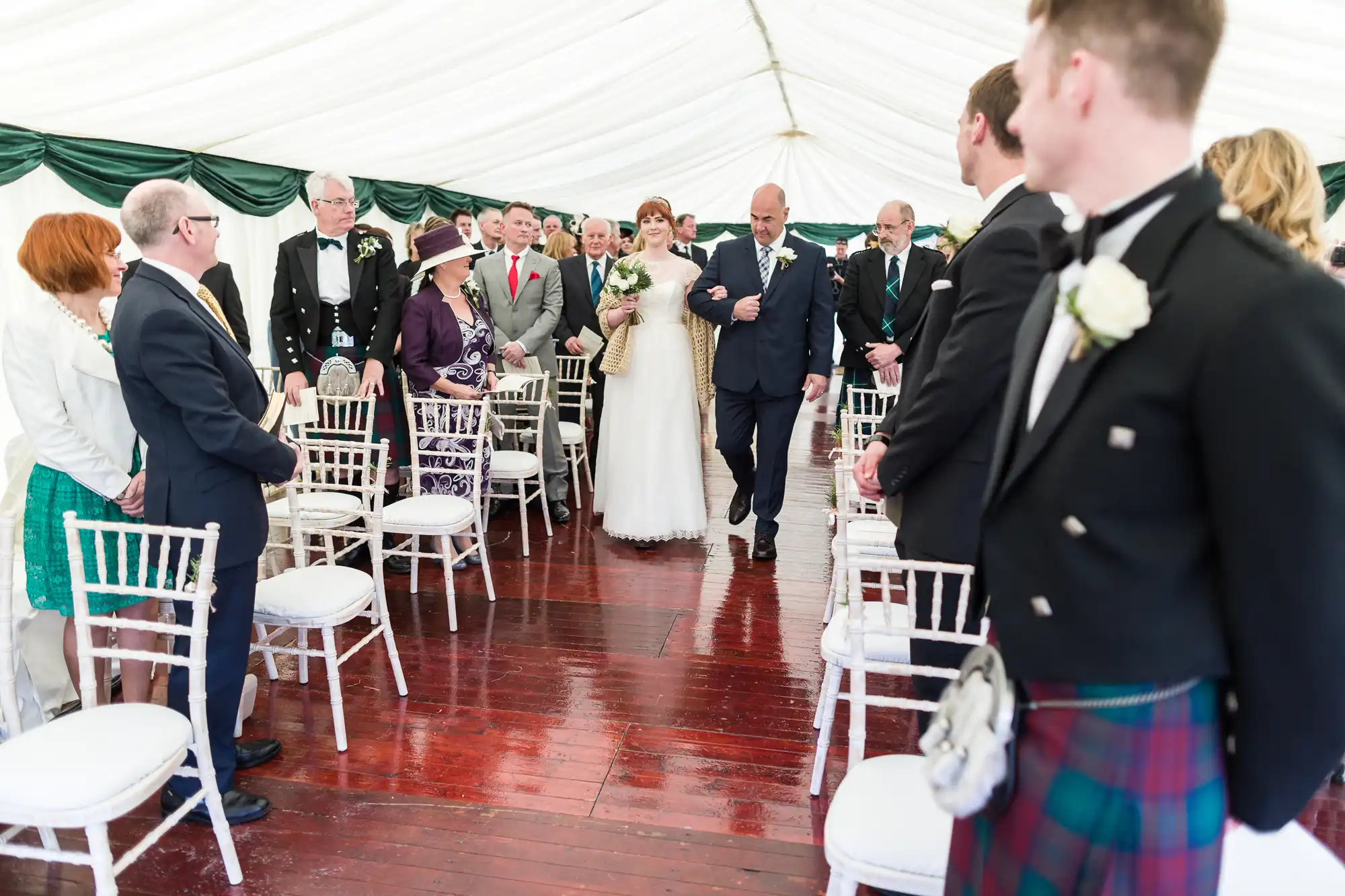 A bride in a white dress holding a bouquet walks down the aisle with an older man, surrounded by seated and standing guests in formal attire inside a white marquee. Some men wear kilts, and guests watch as the bride approaches the altar.