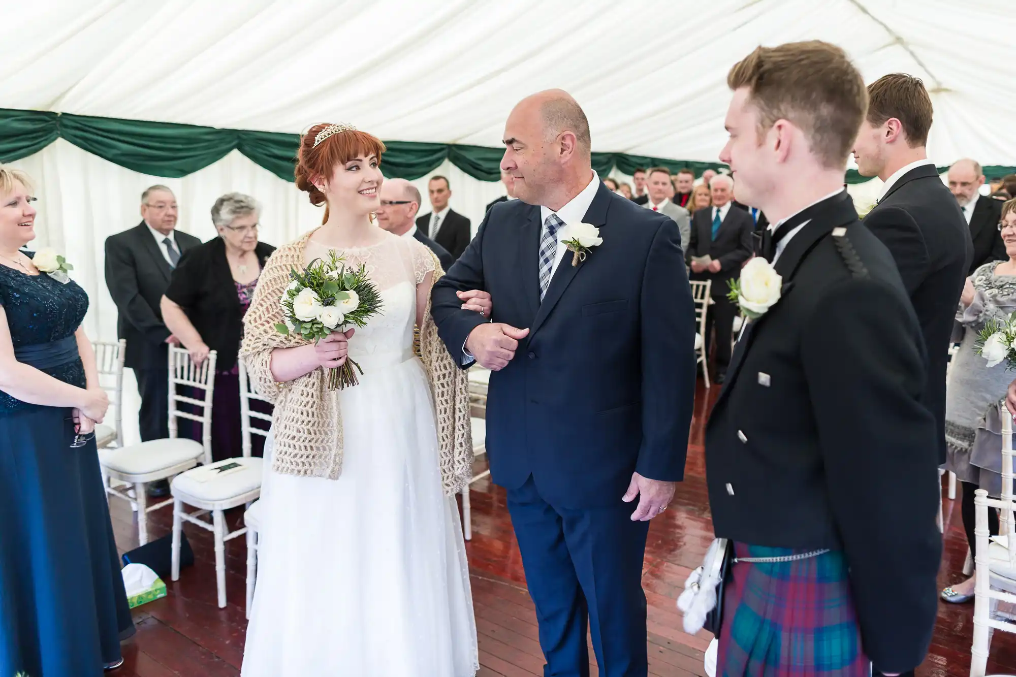 A bride in a white dress and shawl holds a bouquet, smiling as she is escorted down the aisle by an older man in a suit. Guests are seated on white chairs, and a man in a kilt stands at the front. The ceremony is in a white marquee with green drapes.