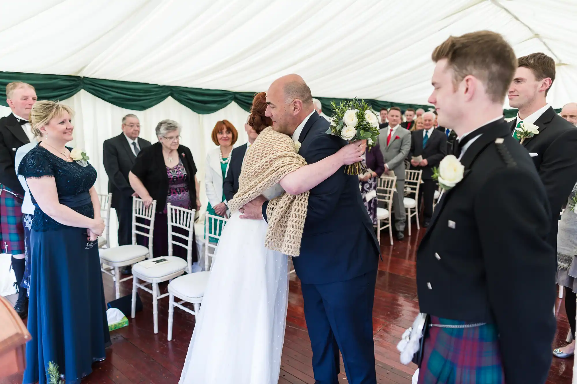 In a white marquee, a bride in a white dress hugs an older man in a blue suit as guests in formal attire, some wearing kilts, watch and smile from white chairs. The mood is joyful and celebratory at this wedding ceremony.