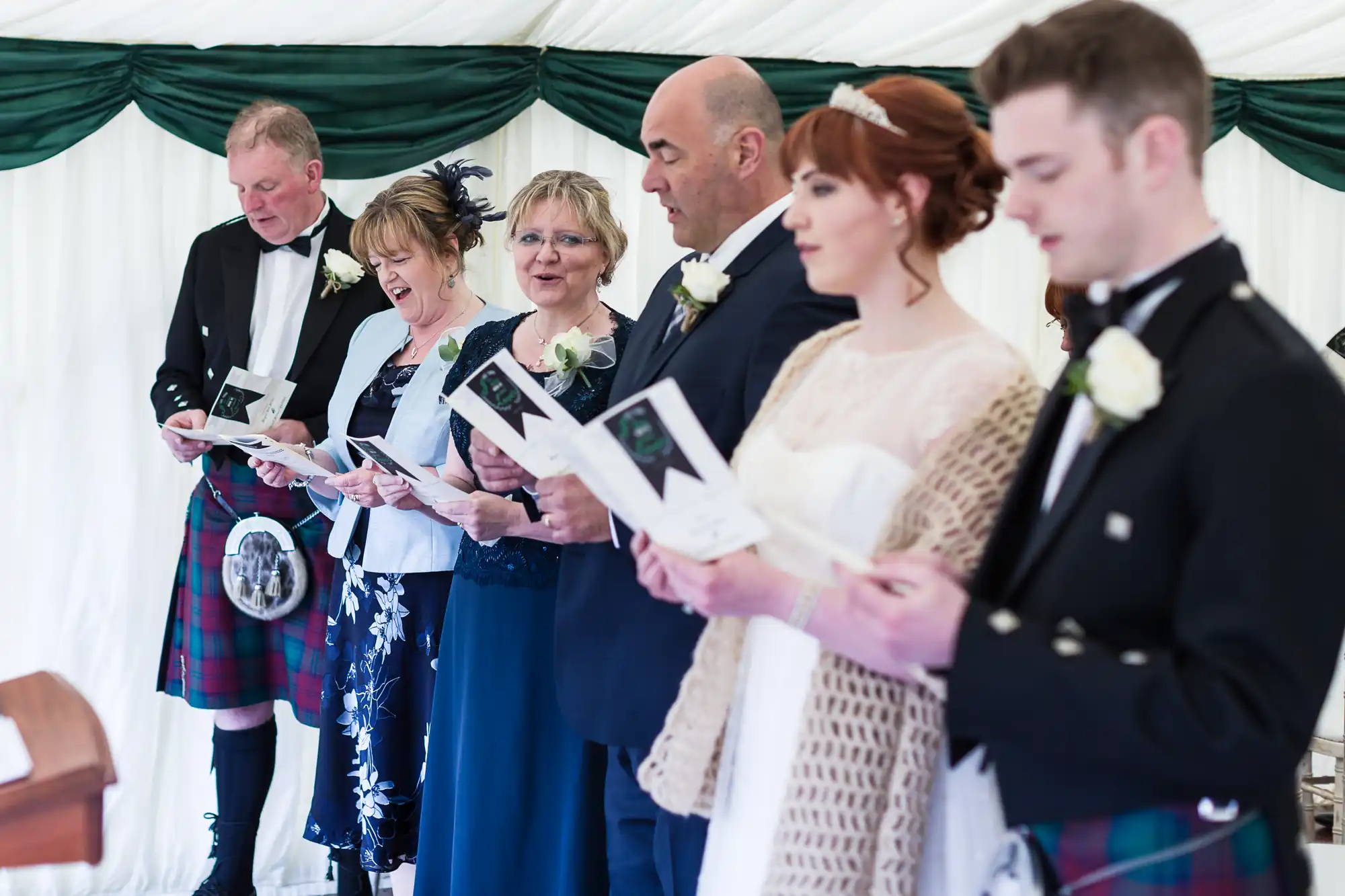 Six people stand in a row indoors, dressed for a formal event. Three men wear kilts and jackets; three women wear dresses. All hold papers and appear to be singing or reading, with white flowers pinned to their outfits. A green and white draped backdrop is behind them.