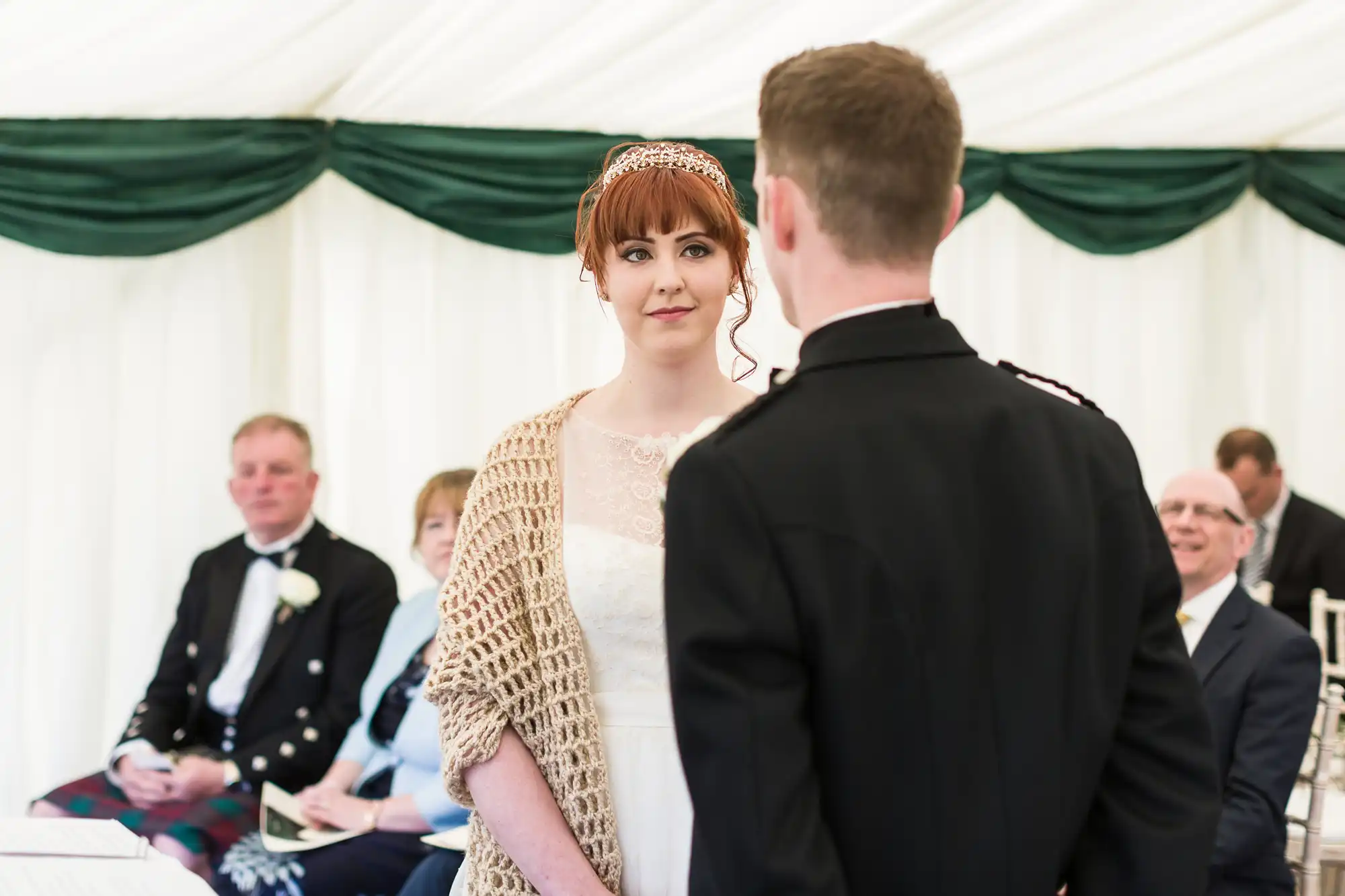 A bride in a white dress and beige shawl faces a groom in a black suit at an indoor wedding ceremony. Both look at each other, while seated guests in formal attire watch in the background, beneath green and white drapery.
