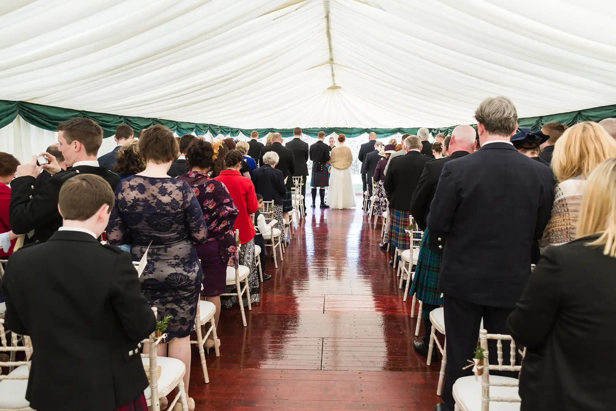 A wedding ceremony inside a white marquee with green trim. Guests in formal attire, including kilts and dresses, stand facing the front where the couple and celebrant are visible. White chairs line both sides of the aisle, and the floor is polished wood.